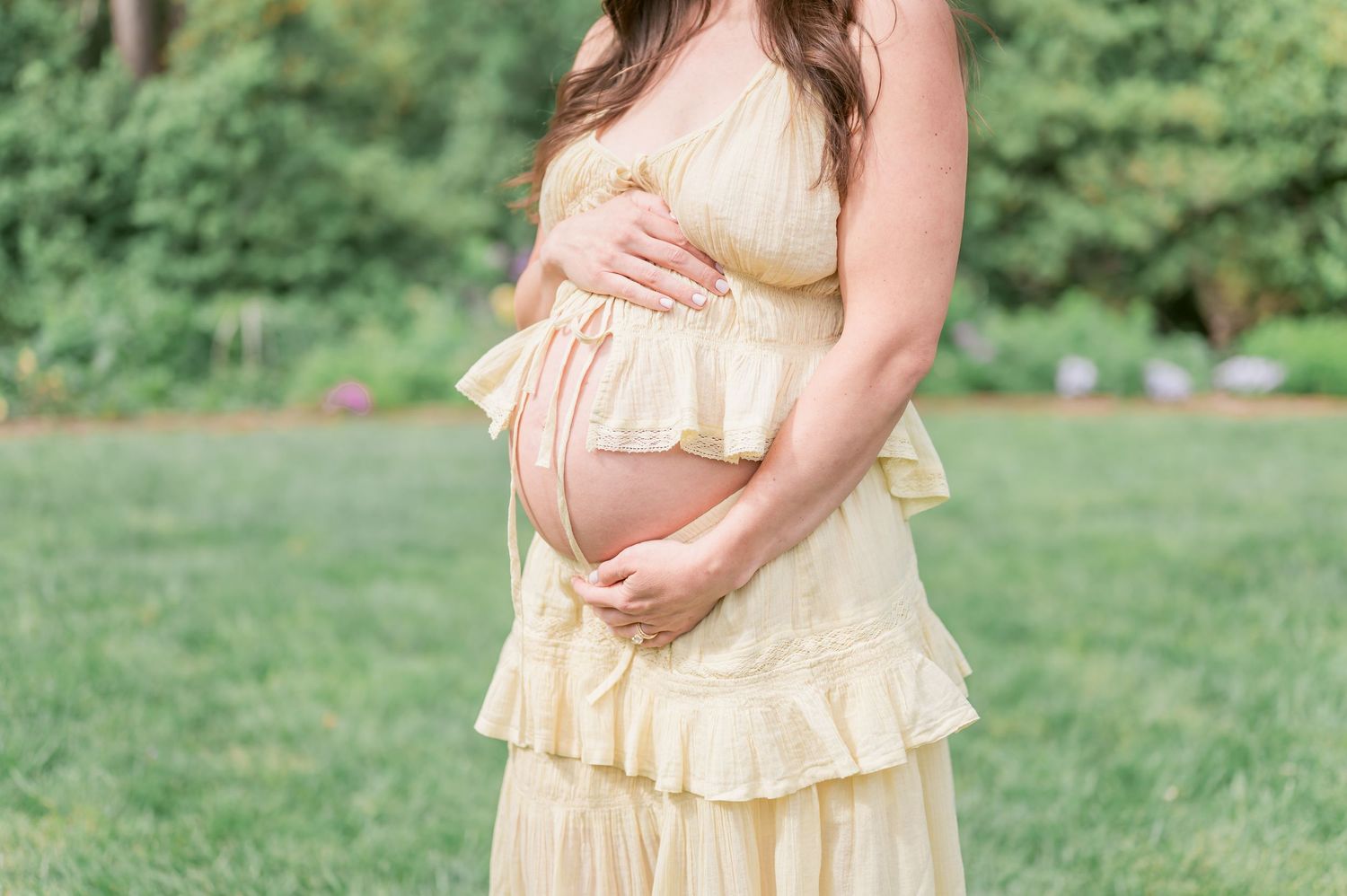 Pregnant woman in yellow ruffled dress cradling baby bump outdoors in lush green garden during maternity photoshoot.
