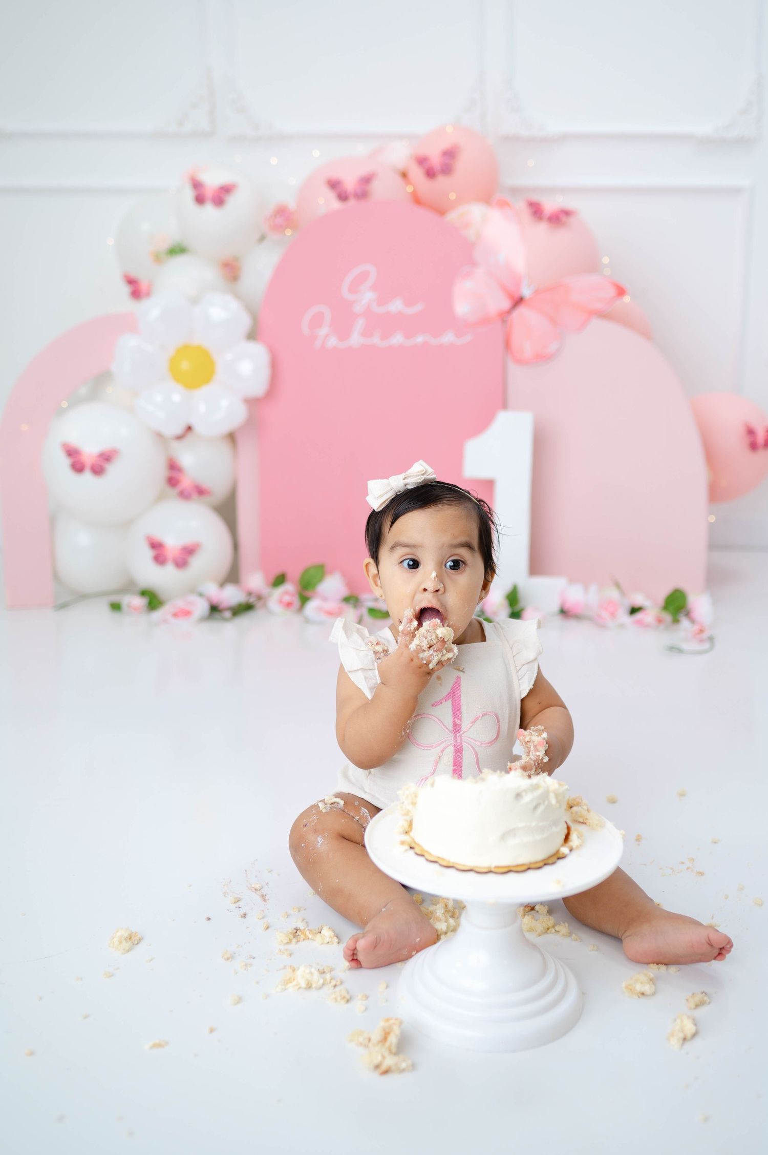 A pink and white themed cake smash photo session with delicate floral decorations and butterflies in the background.