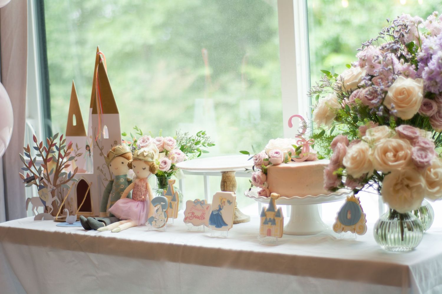 A whimsical party display with a castle centerpiece, delicate floral arrangements, and decorative figurines on a windowsill.