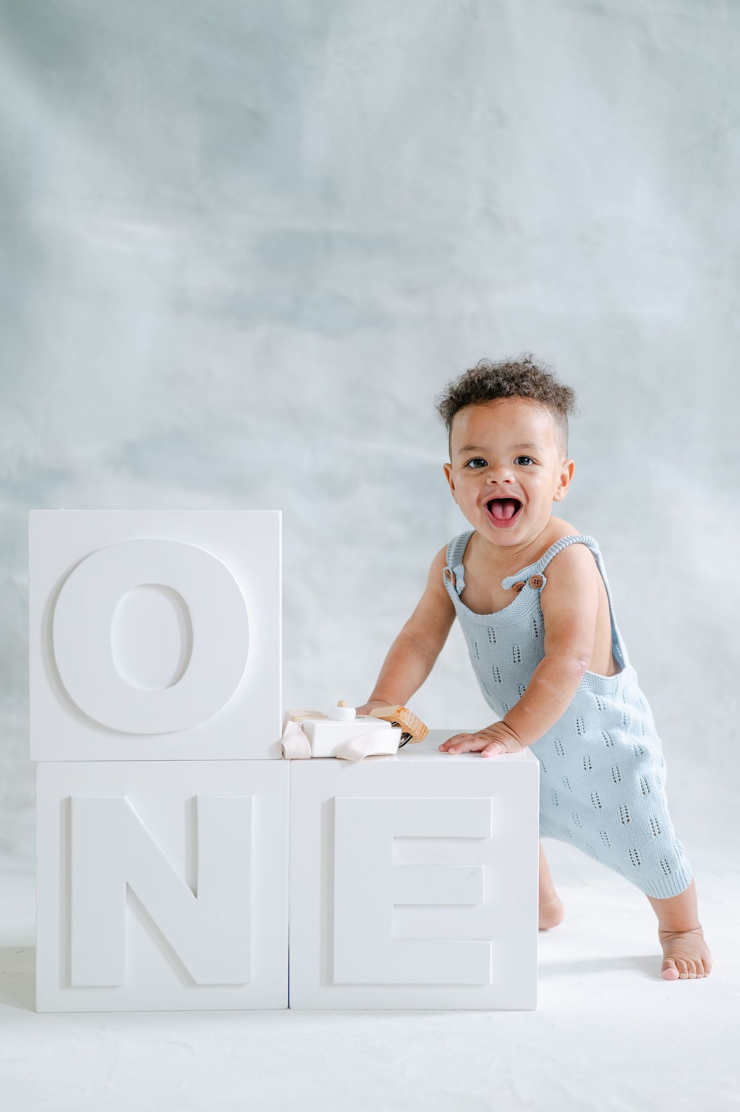 Happy toddler in blue romper standing next to large white ONE letter blocks for first birthday photo.