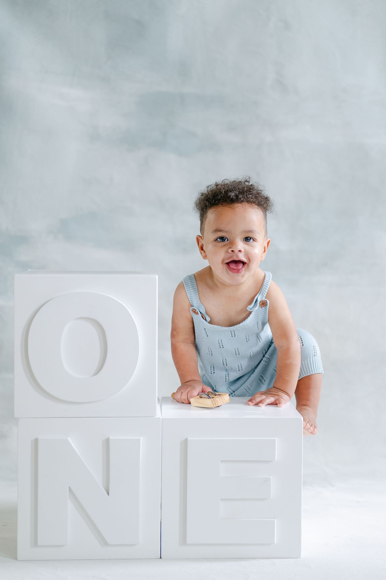 A joyful baby in a blue romper poses with white letter blocks spelling ONE, celebrating their first birthday.