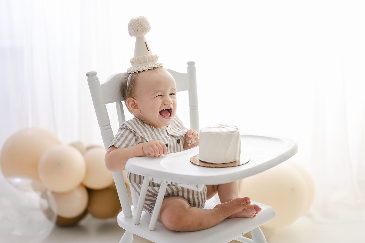 Laughing baby in high chair wearing party hat celebrates first birthday with smash cake and balloons.
