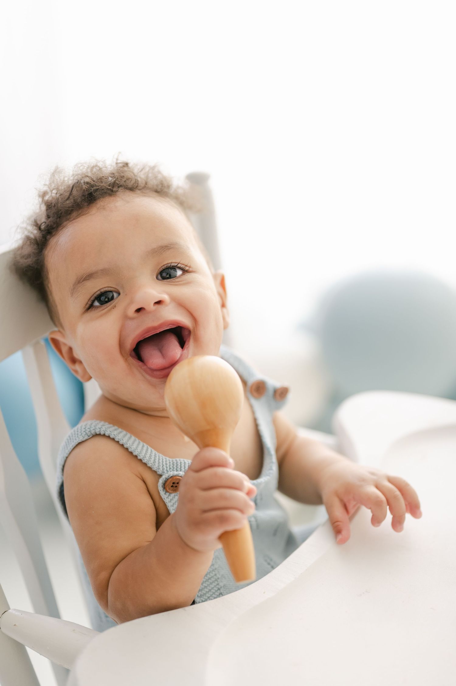 Happy baby in high chair holding wooden spoon toy with mouth open, wearing gray romper in bright white room.