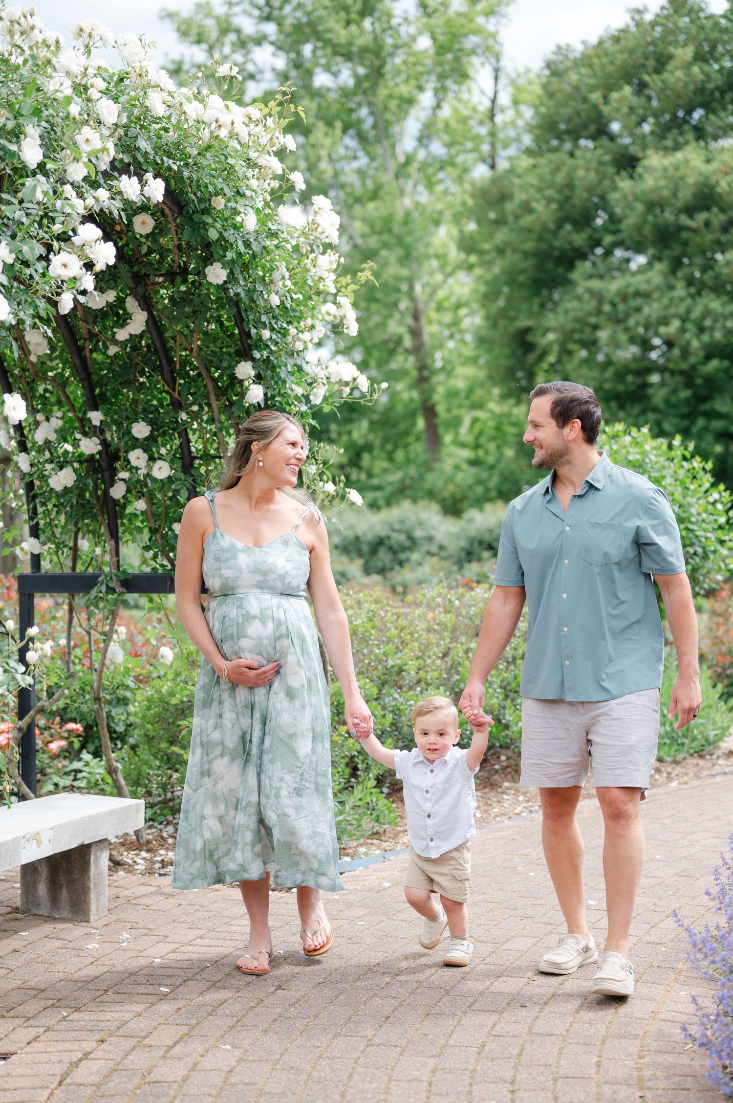 Pregnant woman in floral dress walks with toddler and man in garden with white roses blooming overhead.