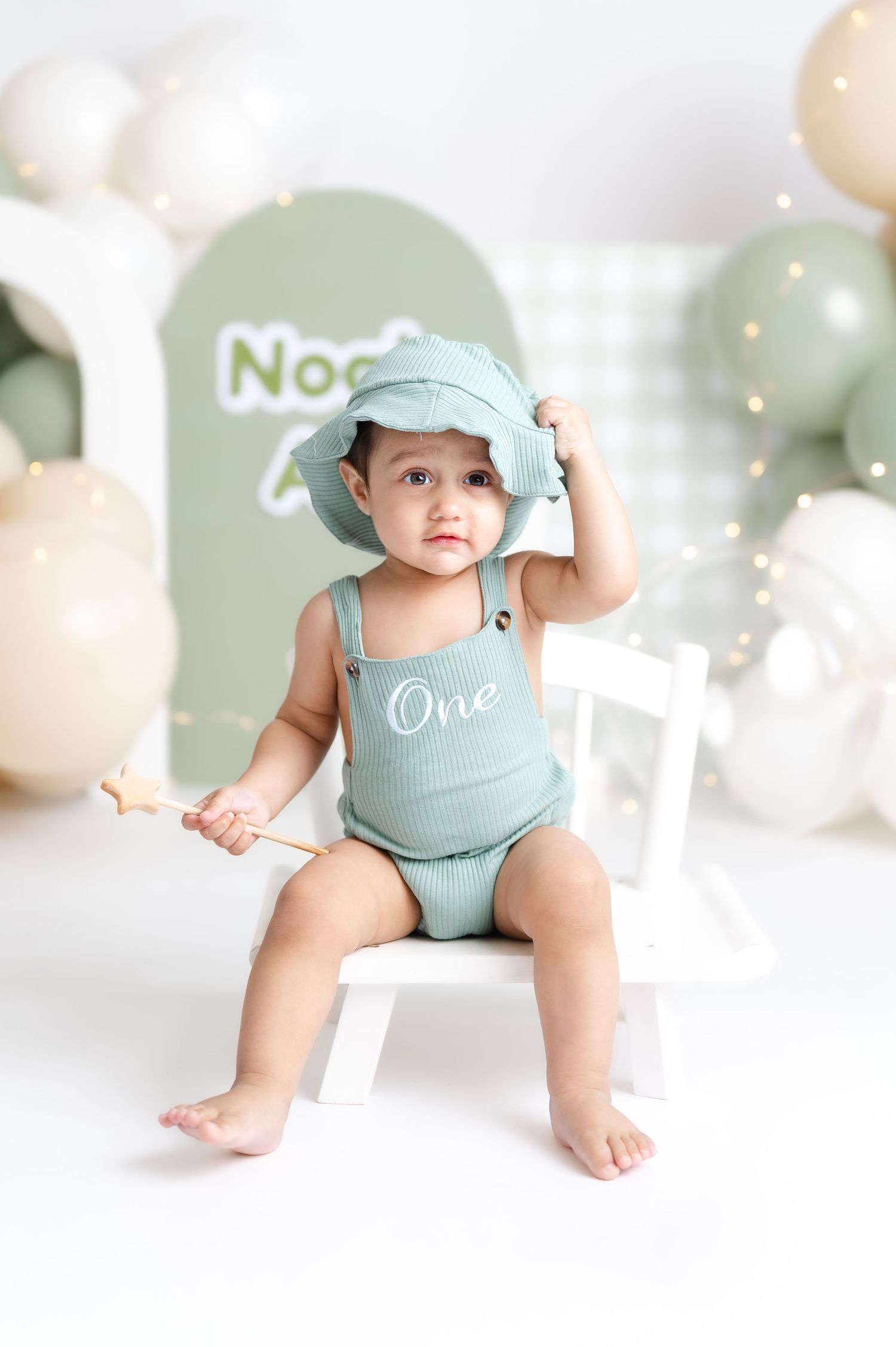 A baby in a green romper and bucket hat sits on a white chair holding a star wand at a first birthday photo shoot.