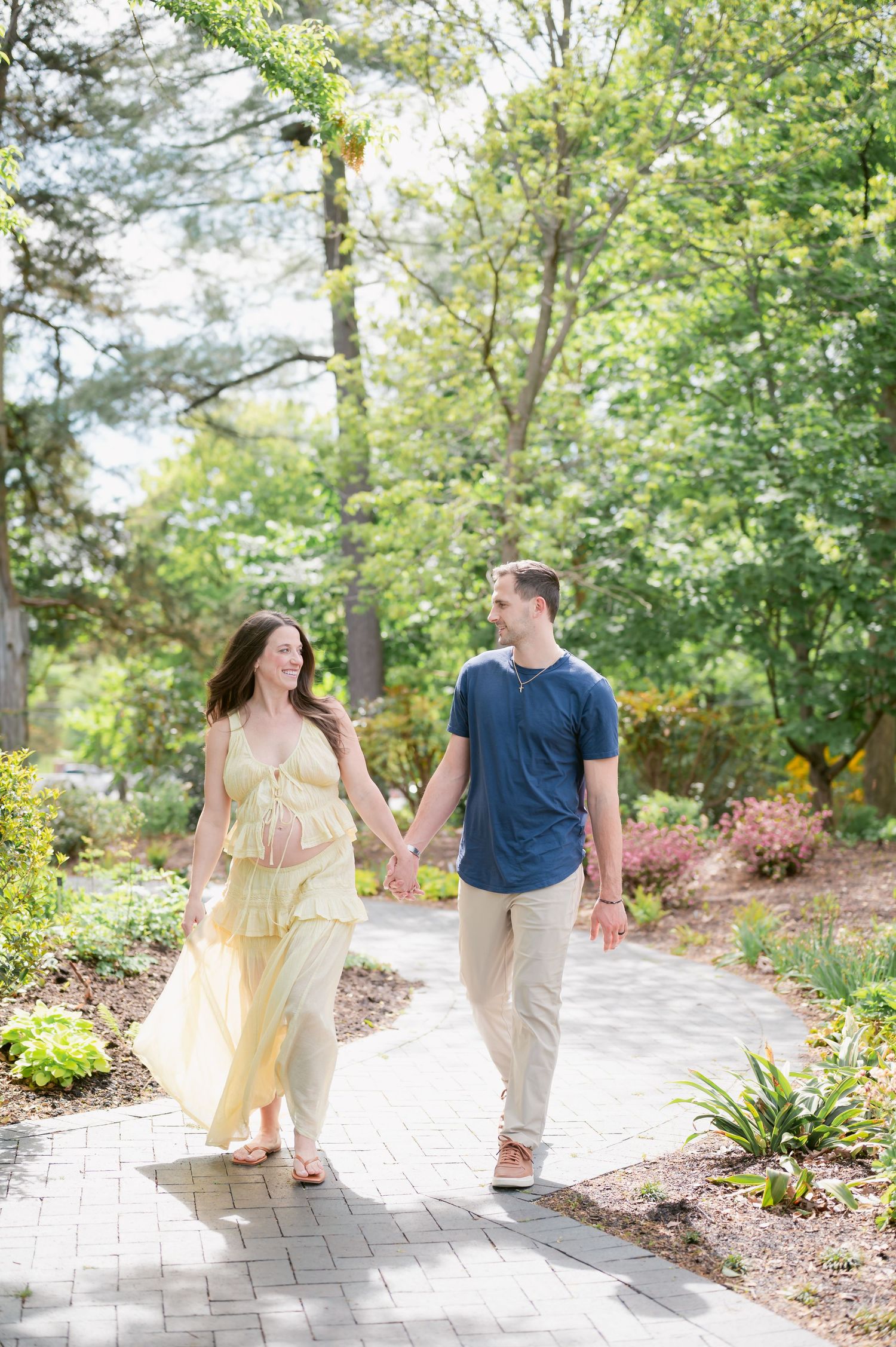 Pregnant couple holding hands walking on a garden path surrounded by lush green trees during a maternity photoshoot.