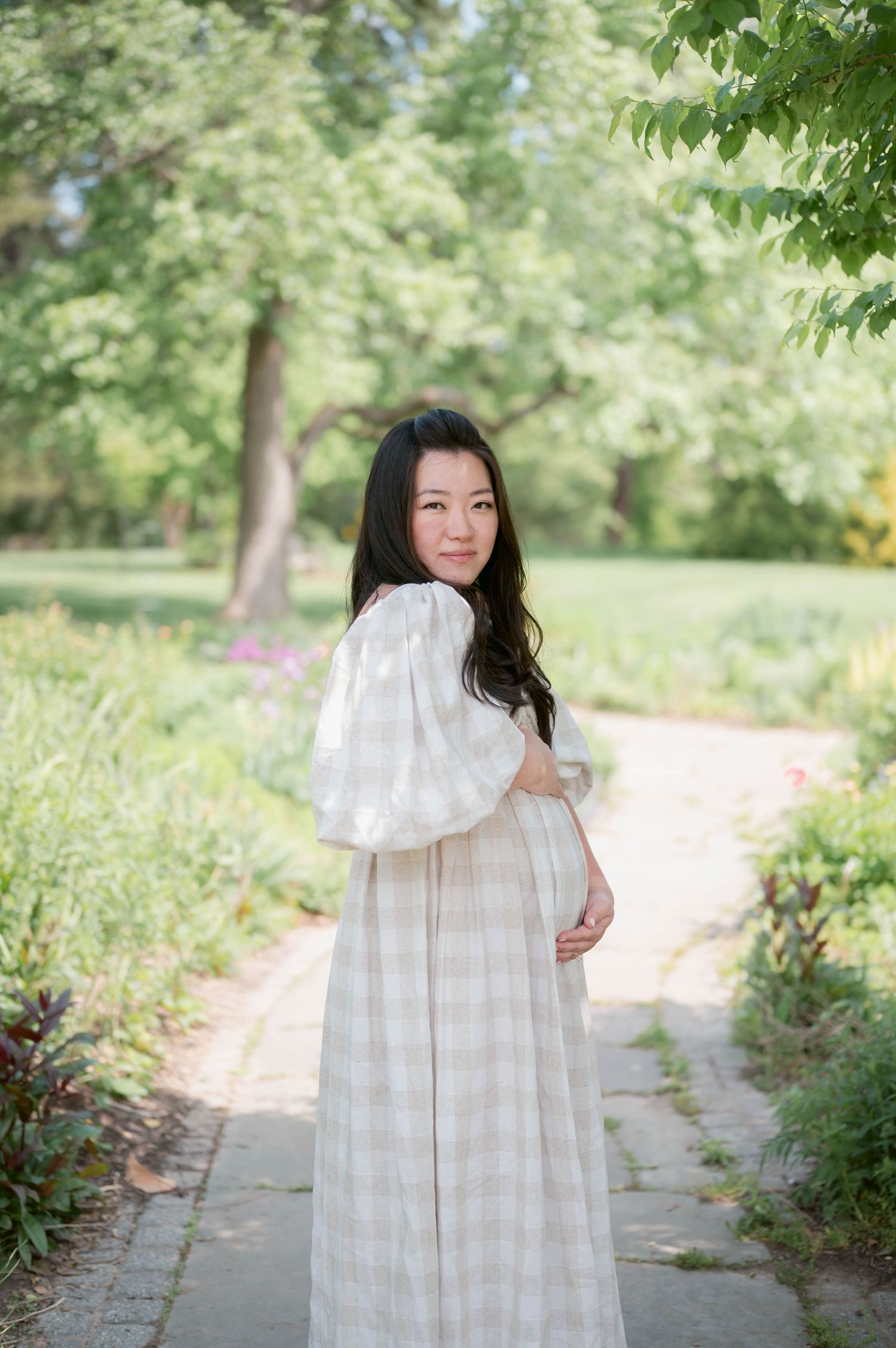 Pregnant woman in white plaid dress holding baby bump on garden path surrounded by lush green trees.