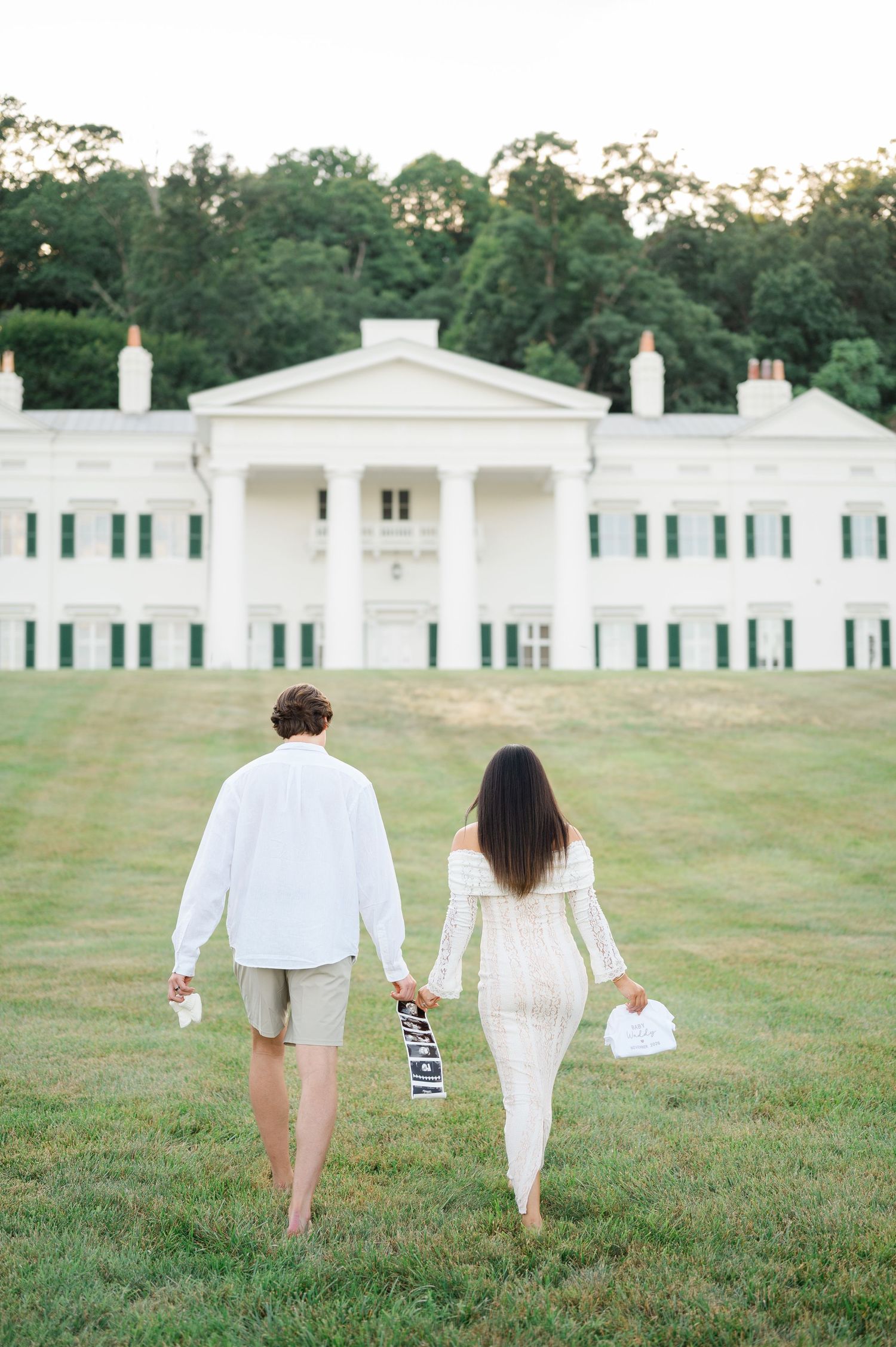 Couple walks hand-in-hand toward a grand white mansion with green shutters and columns on a lush lawn.