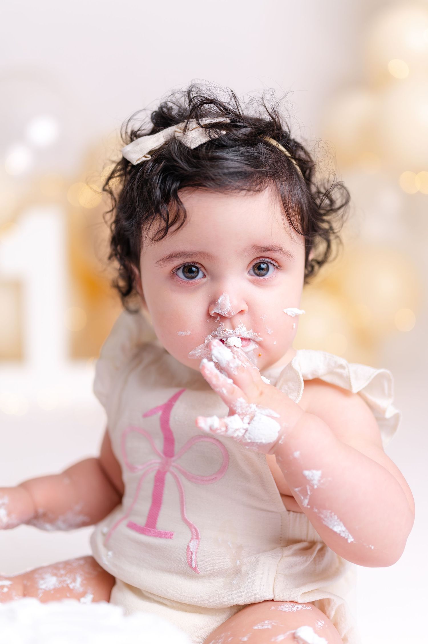 Baby girl with cake smash on her face celebrating her first birthday, wearing a cream romper with a pink number one.