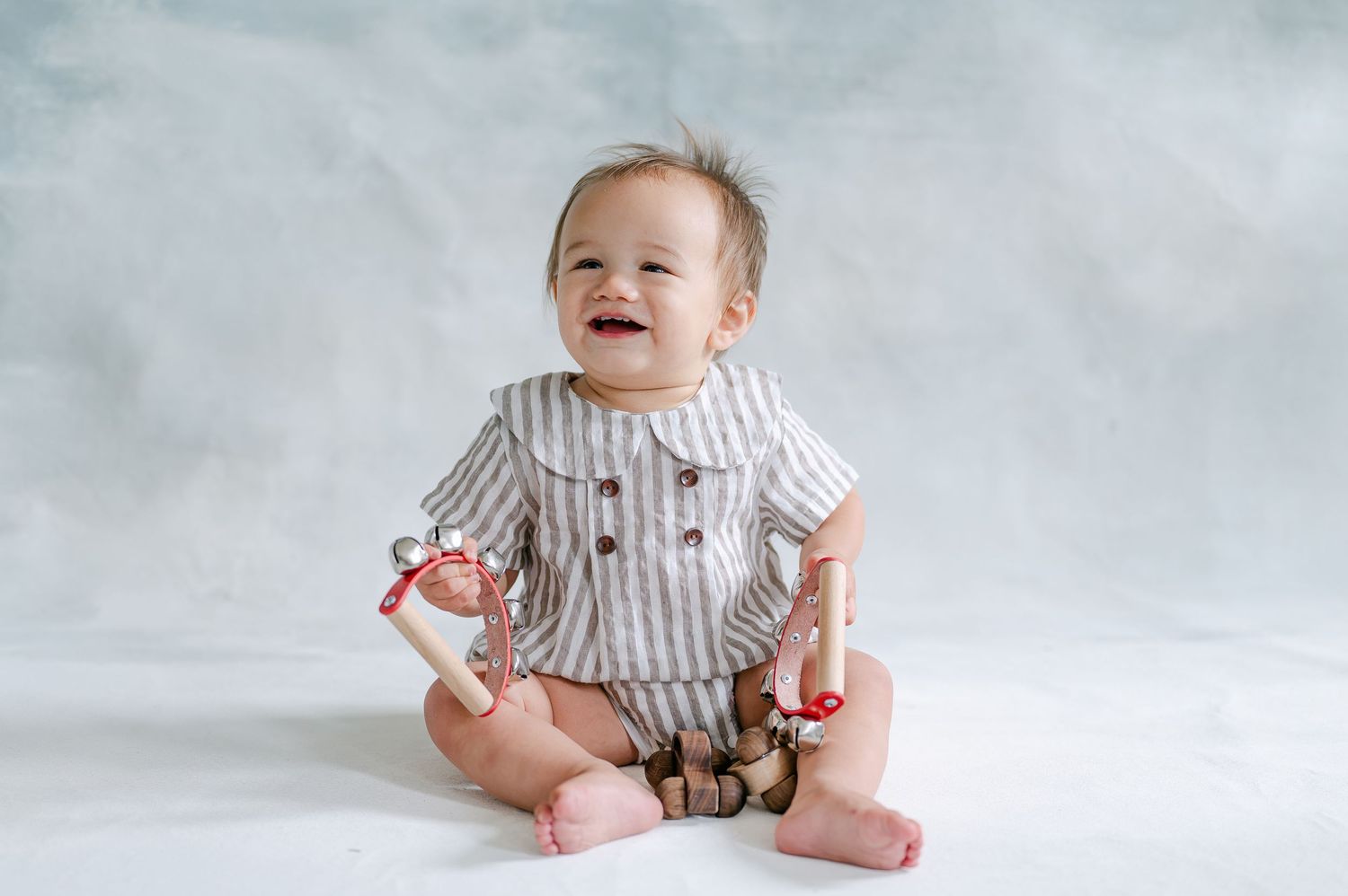 Smiling baby in striped romper sitting on white backdrop, holding wooden toy rattles with red ribbons.
