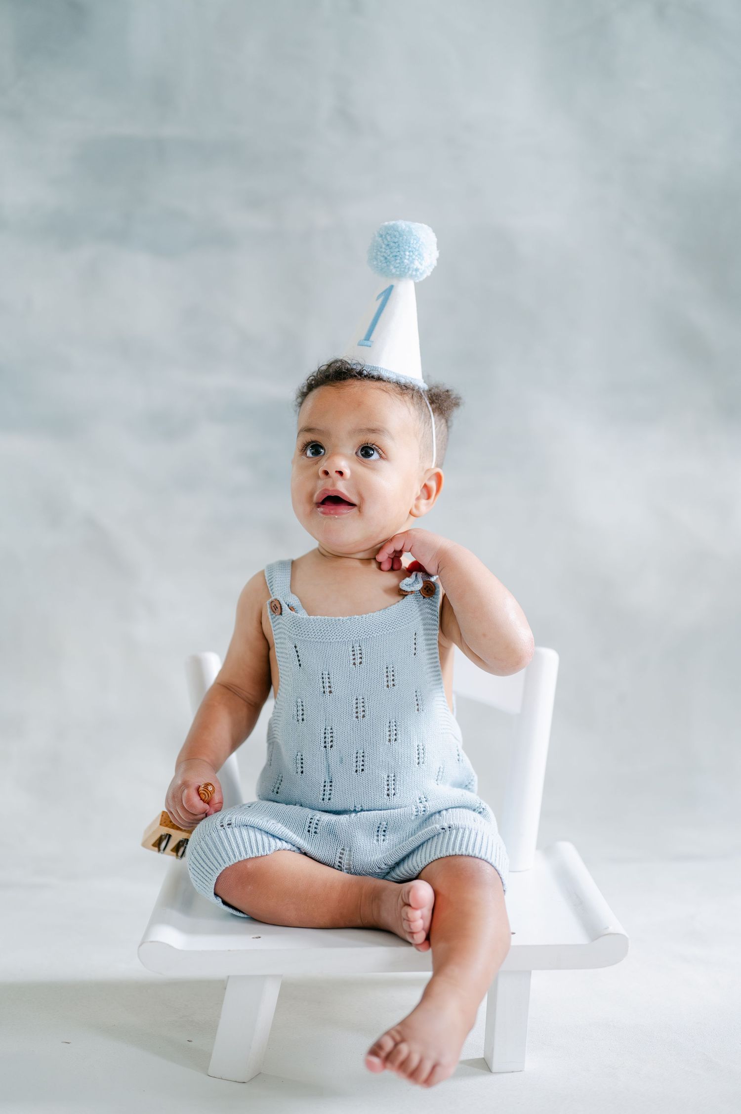A baby in a light blue knit romper and birthday party hat sits on a white chair, looking up curiously.