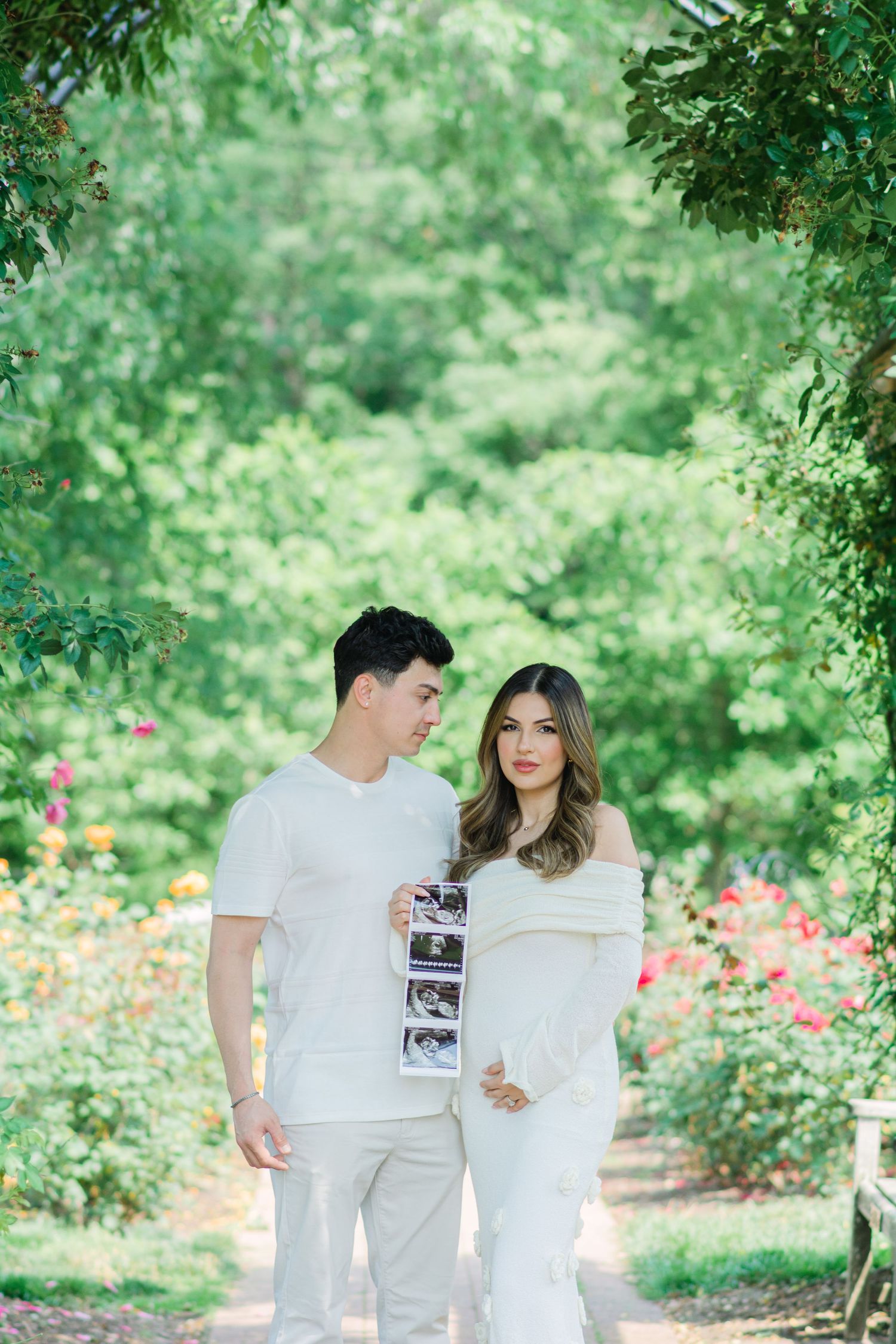 Romantic pregnancy announcement photoshoot in a garden with white outfits and ultrasound photos.