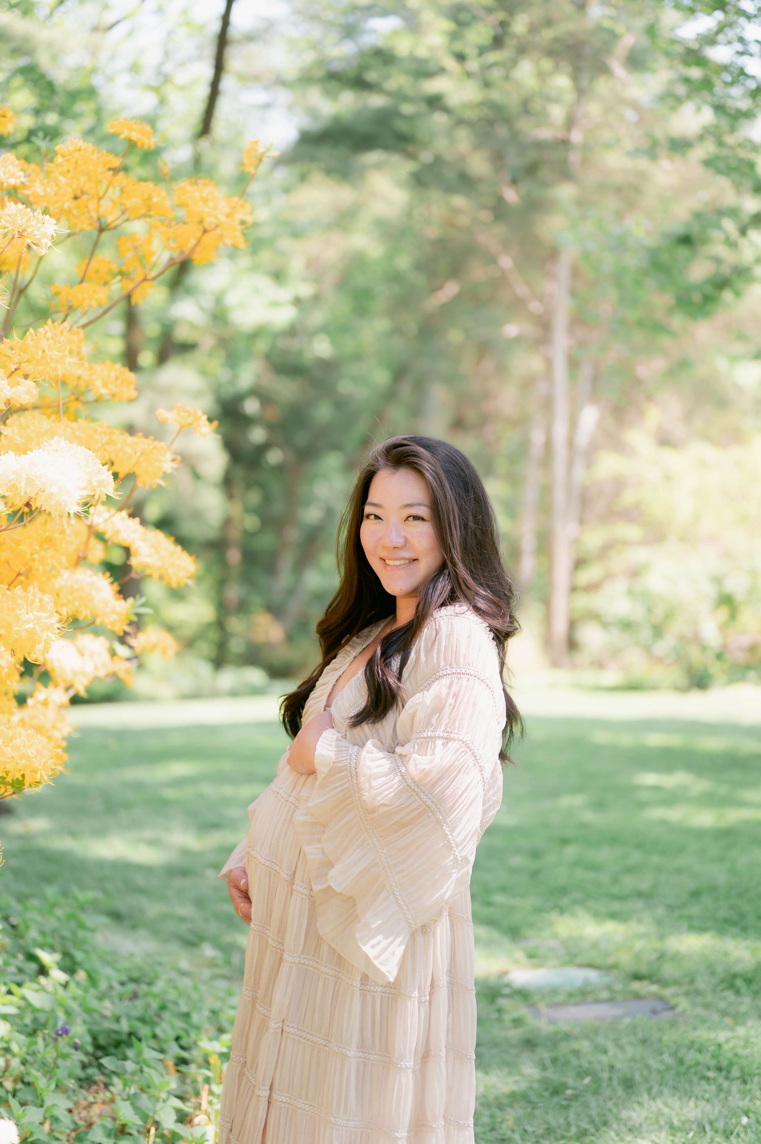 Smiling pregnant woman in cream dress cradling baby bump outdoors near yellow flowers in lush green garden.