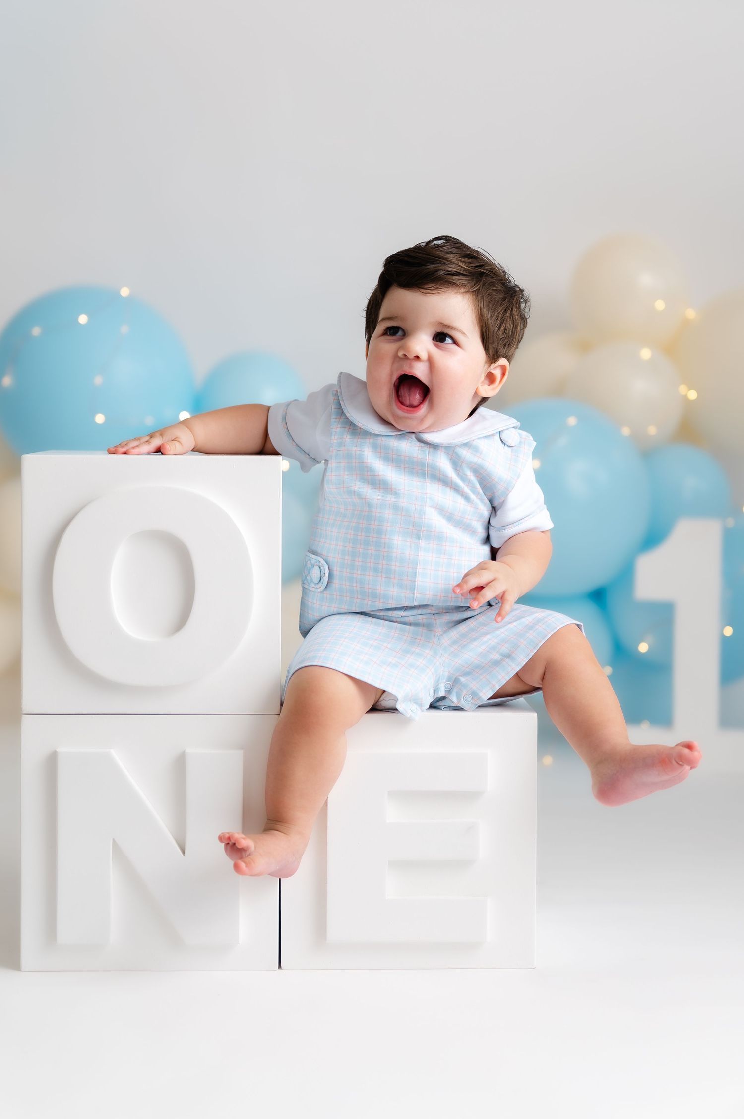 Baby boy in blue outfit sits on white ONE blocks with balloons for his first birthday photo shoot.