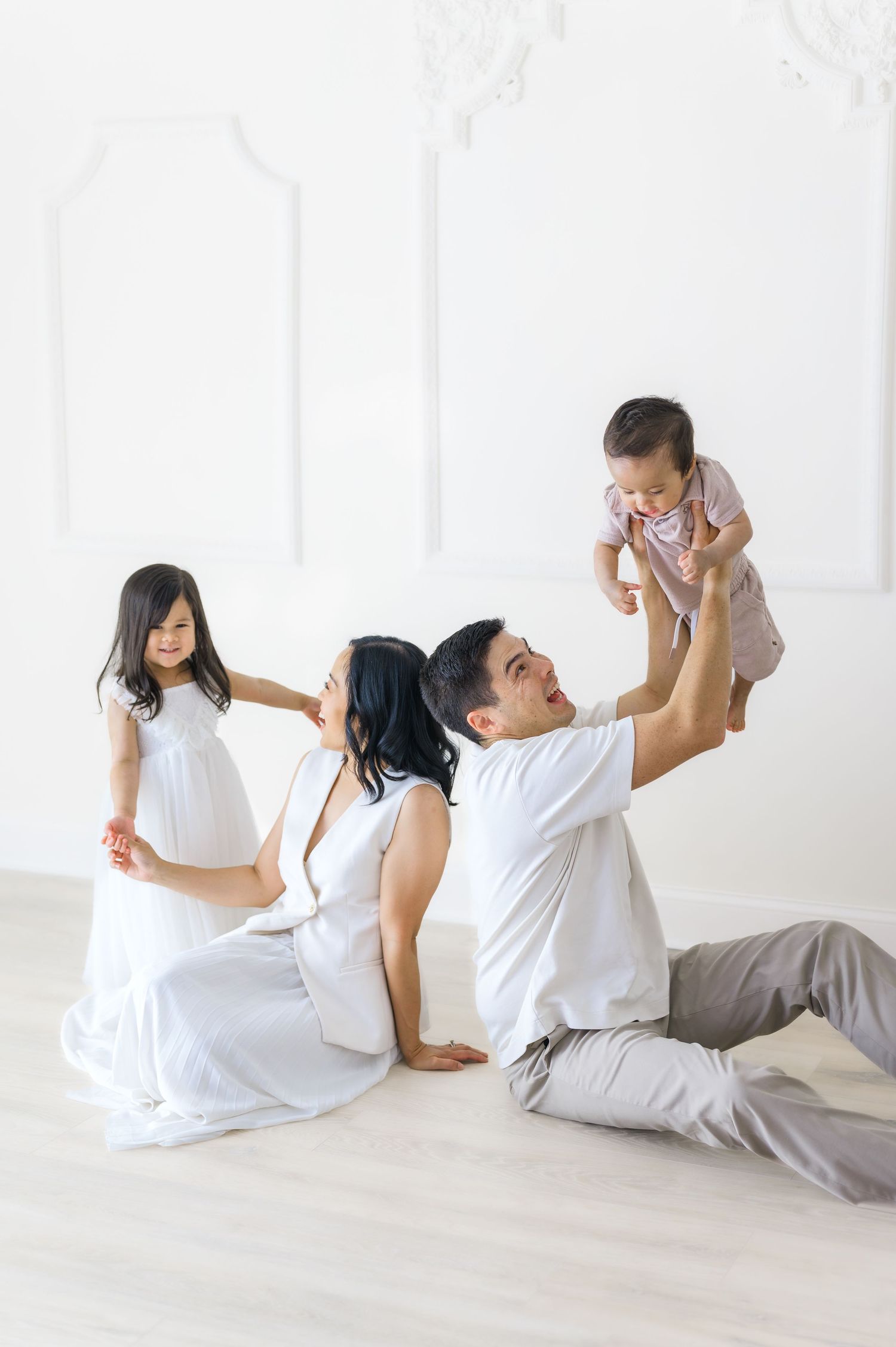 Happy Asian family playing together on white floor, father lifting baby while mother and daughter smile joyfully.