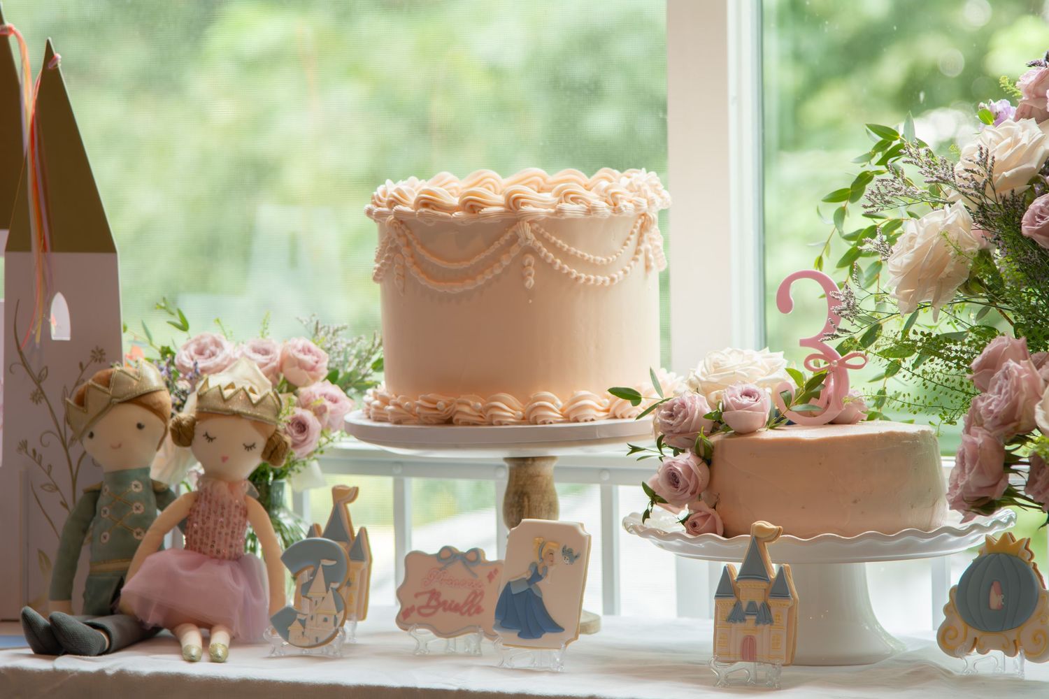 A pale pink ruffled cake sits on a white stand surrounded by soft floral arrangements and decorative figurines.