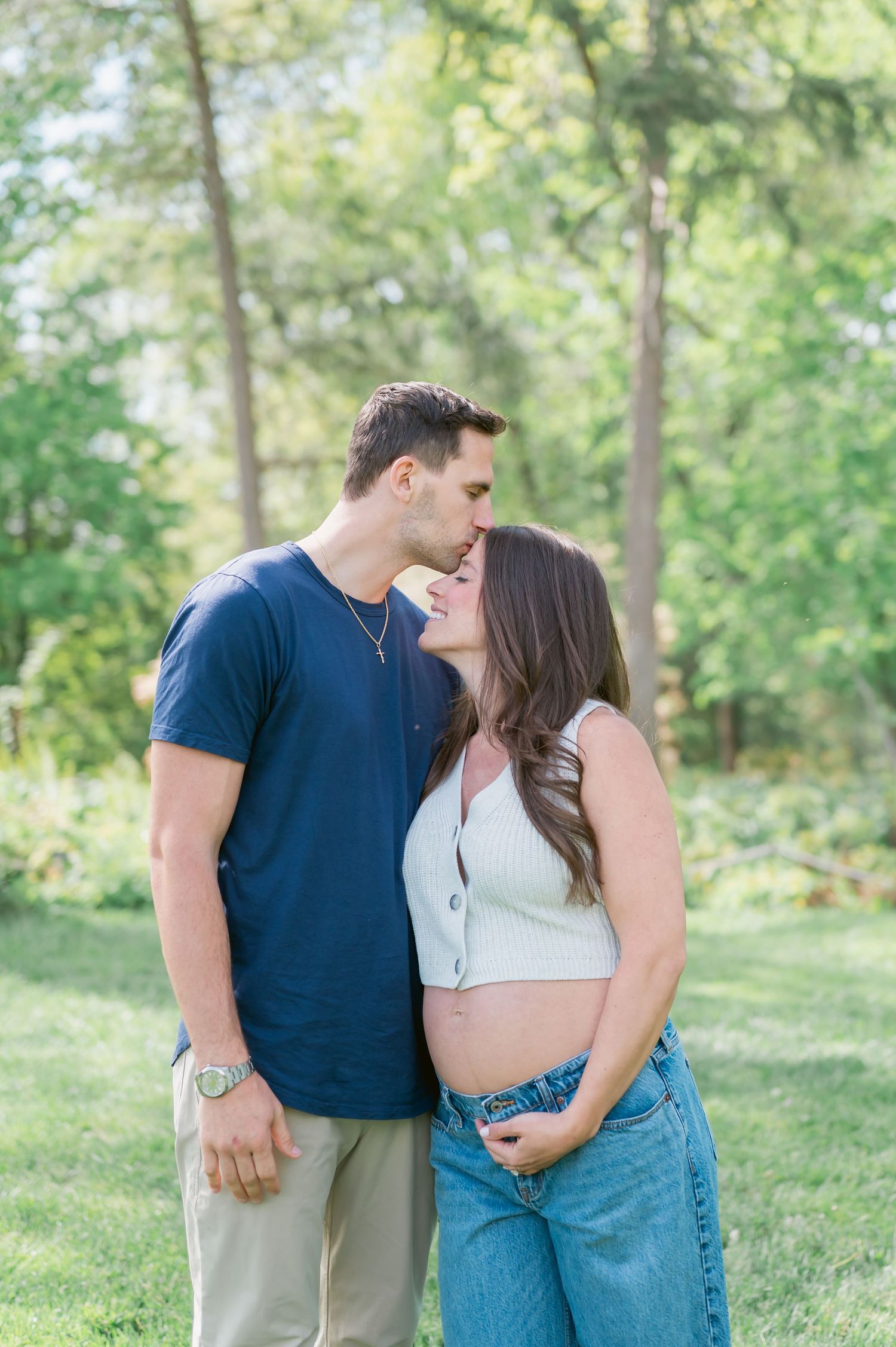 Couple sharing a tender kiss outdoors, woman showing baby bump in white crop top and jeans, man in navy shirt, lush green trees background.