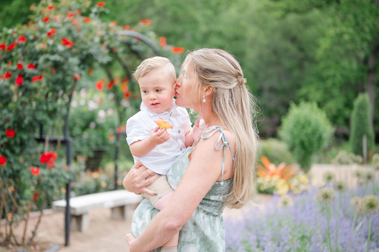 Mother kissing baby boy in garden with red roses and lavender flowers in soft natural light.