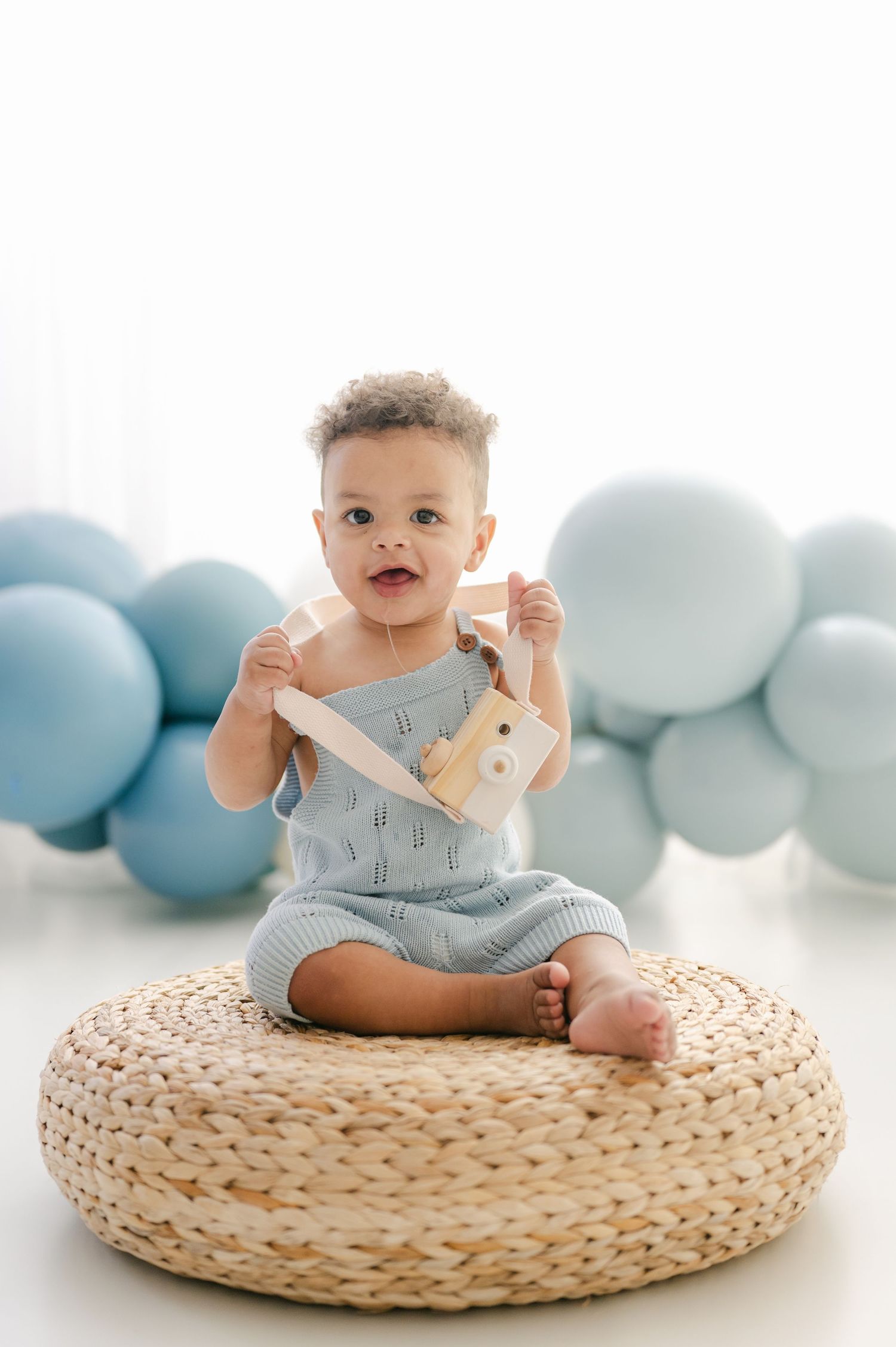Smiling baby boy in blue romper sits on wicker pouf holding wooden toy camera surrounded by blue balloons.