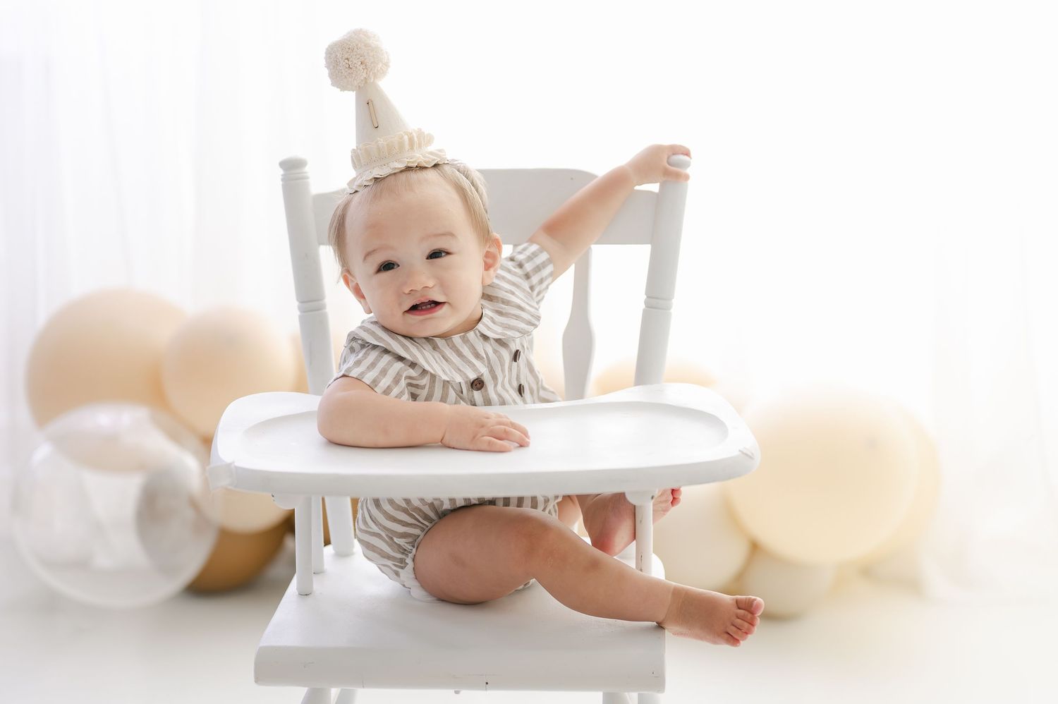 A smiling baby in a striped outfit and party hat sits in a white high chair surrounded by neutral balloons.