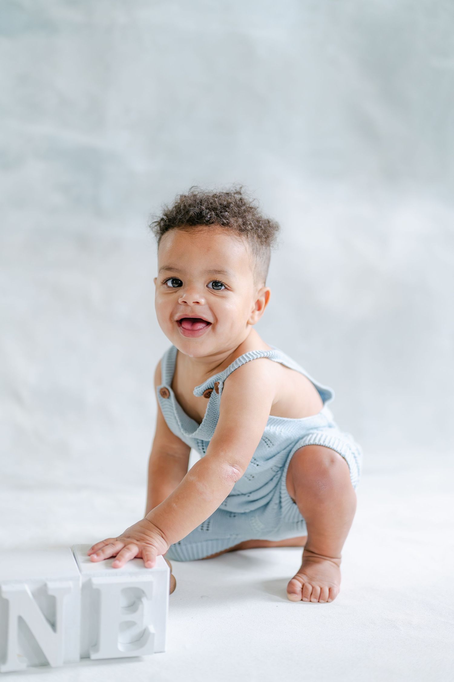 A smiling baby in a blue knit romper sits next to letter blocks spelling ONE, celebrating their first birthday.