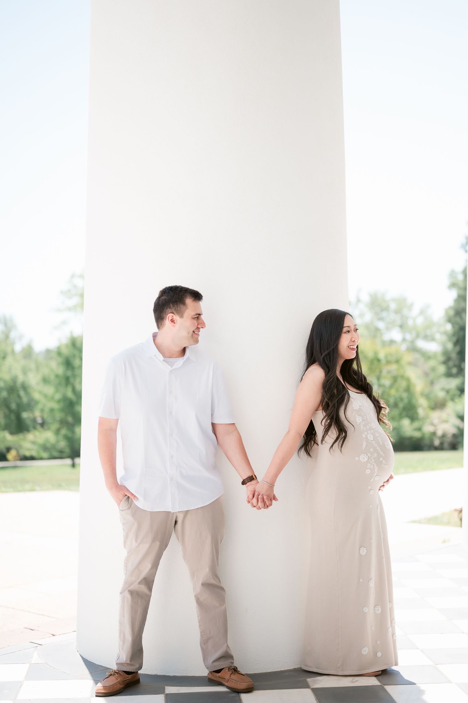 Expecting couple holds hands smiling at each other leaning against white column outdoors in matching neutral outfits.