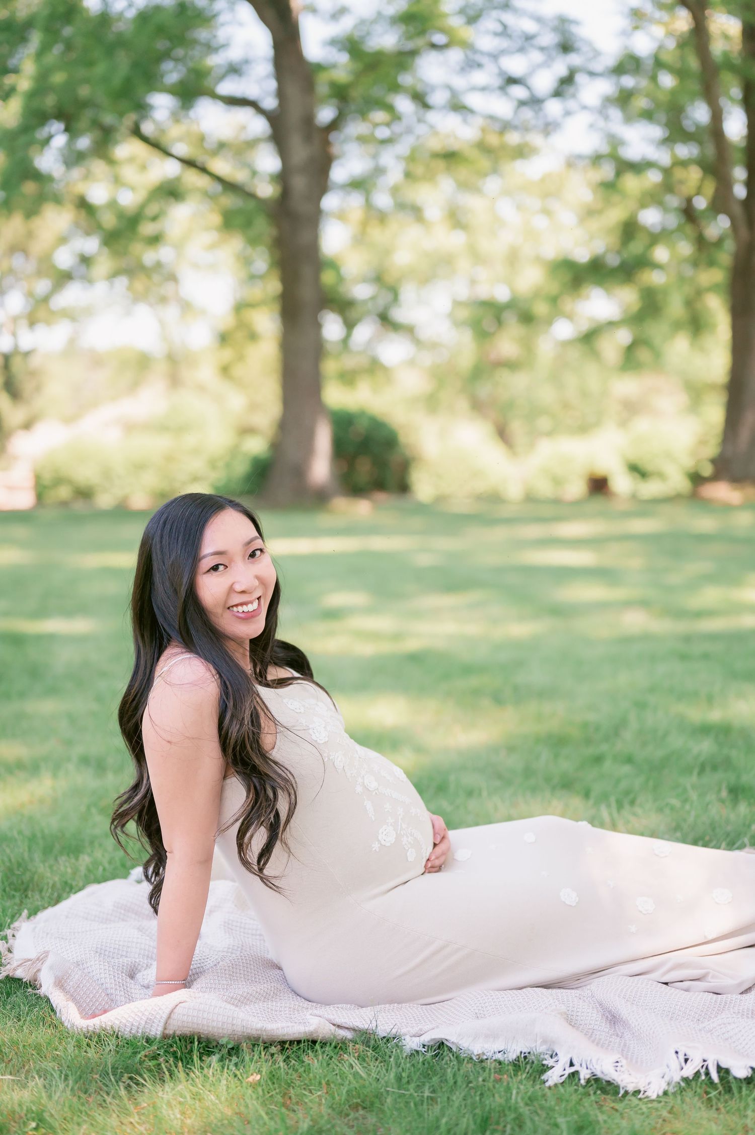 Smiling pregnant woman in cream dress sitting on blanket in lush green park during outdoor maternity photo session.