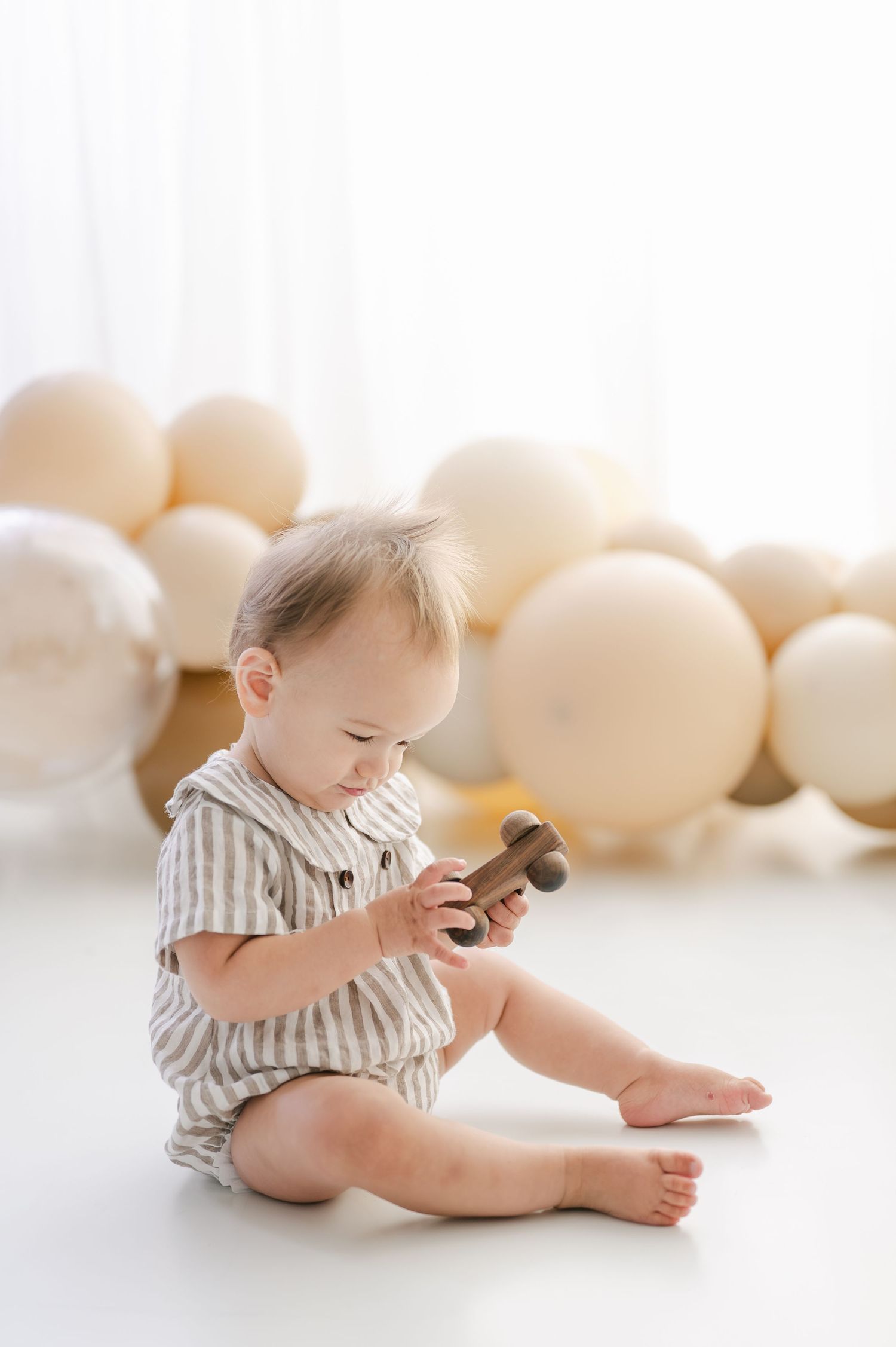 A baby in a striped romper sits on a white floor playing with a wooden toy, surrounded by cream balloons.
