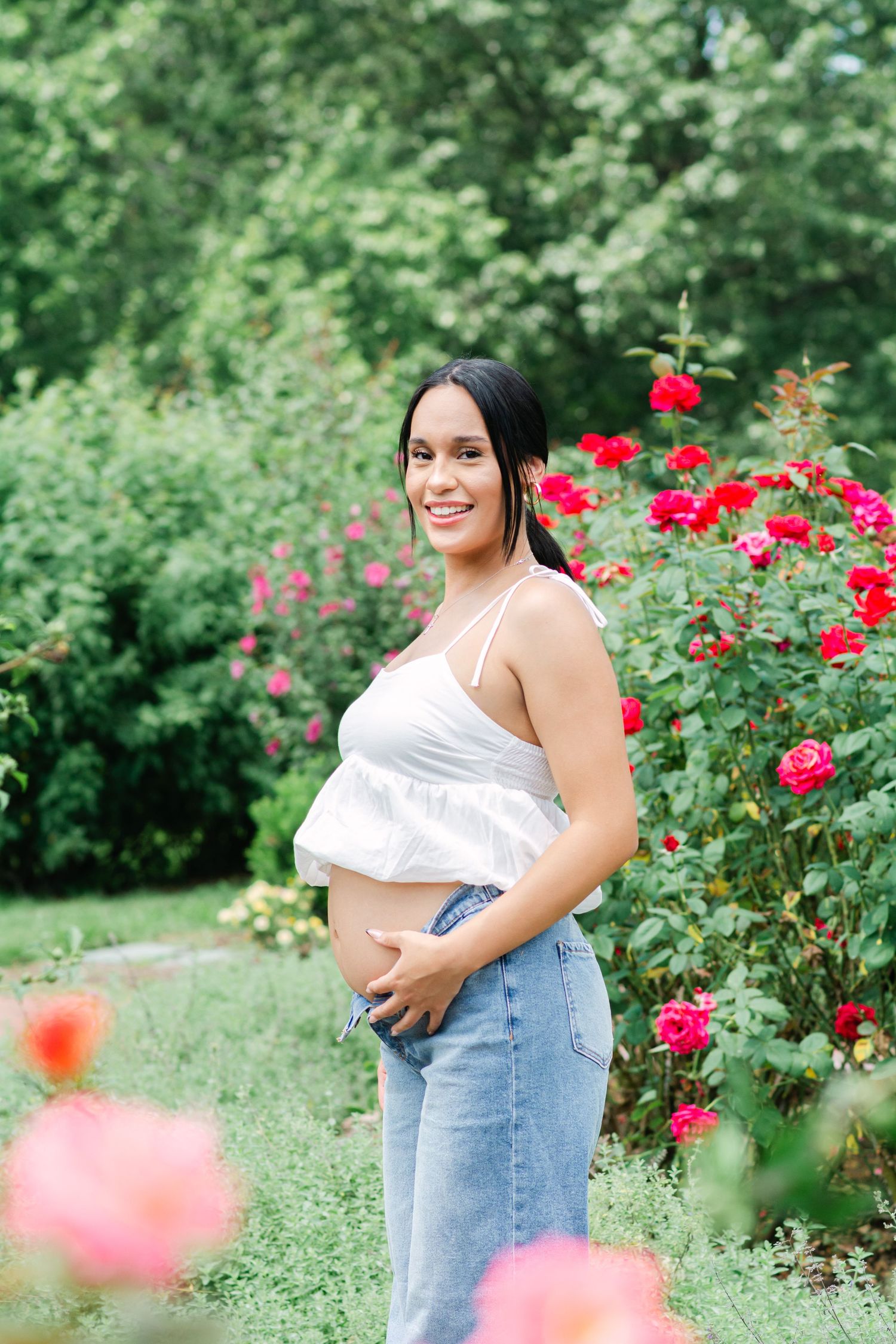 Outdoor maternity photoshoot with red roses and lush greenery in the background.