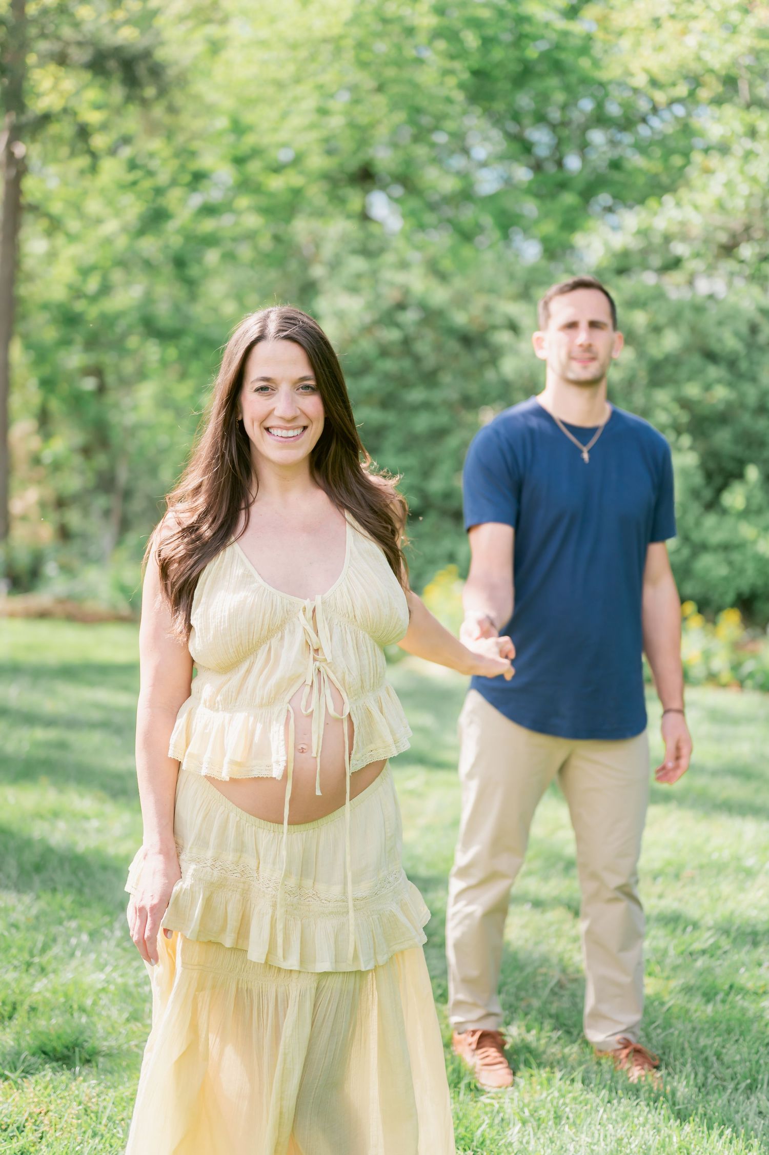 Pregnant woman in yellow dress holds hands with man in navy shirt during outdoor maternity photoshoot in green park.
