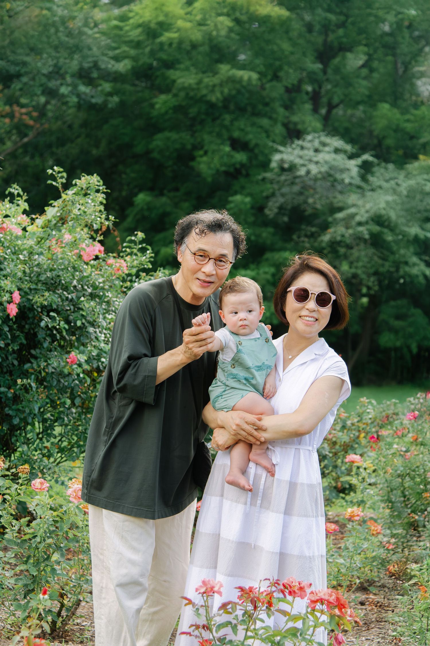 Family photo session in a rose garden with natural sunlight filtering through trees.