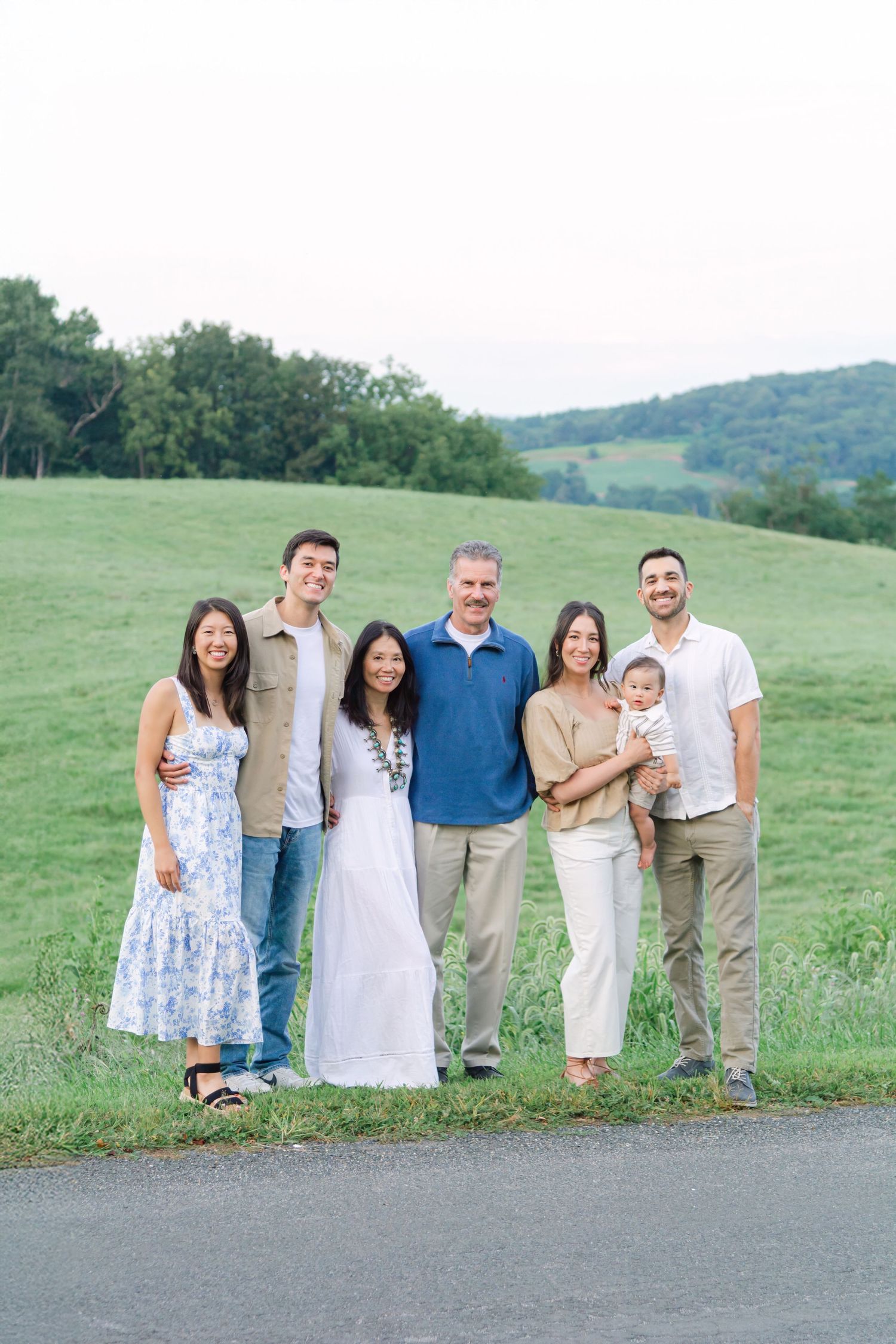 A multi-generational family portrait outdoors in a pastoral setting with rolling hills and fields in the background.