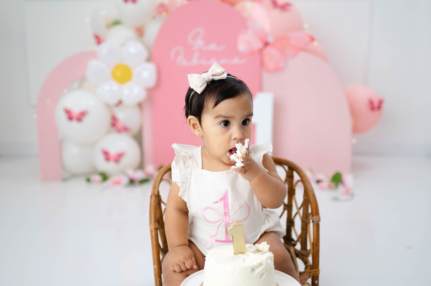 Baby in white dress sits in wooden high chair with pink and floral backdrop for first birthday photoshoot.