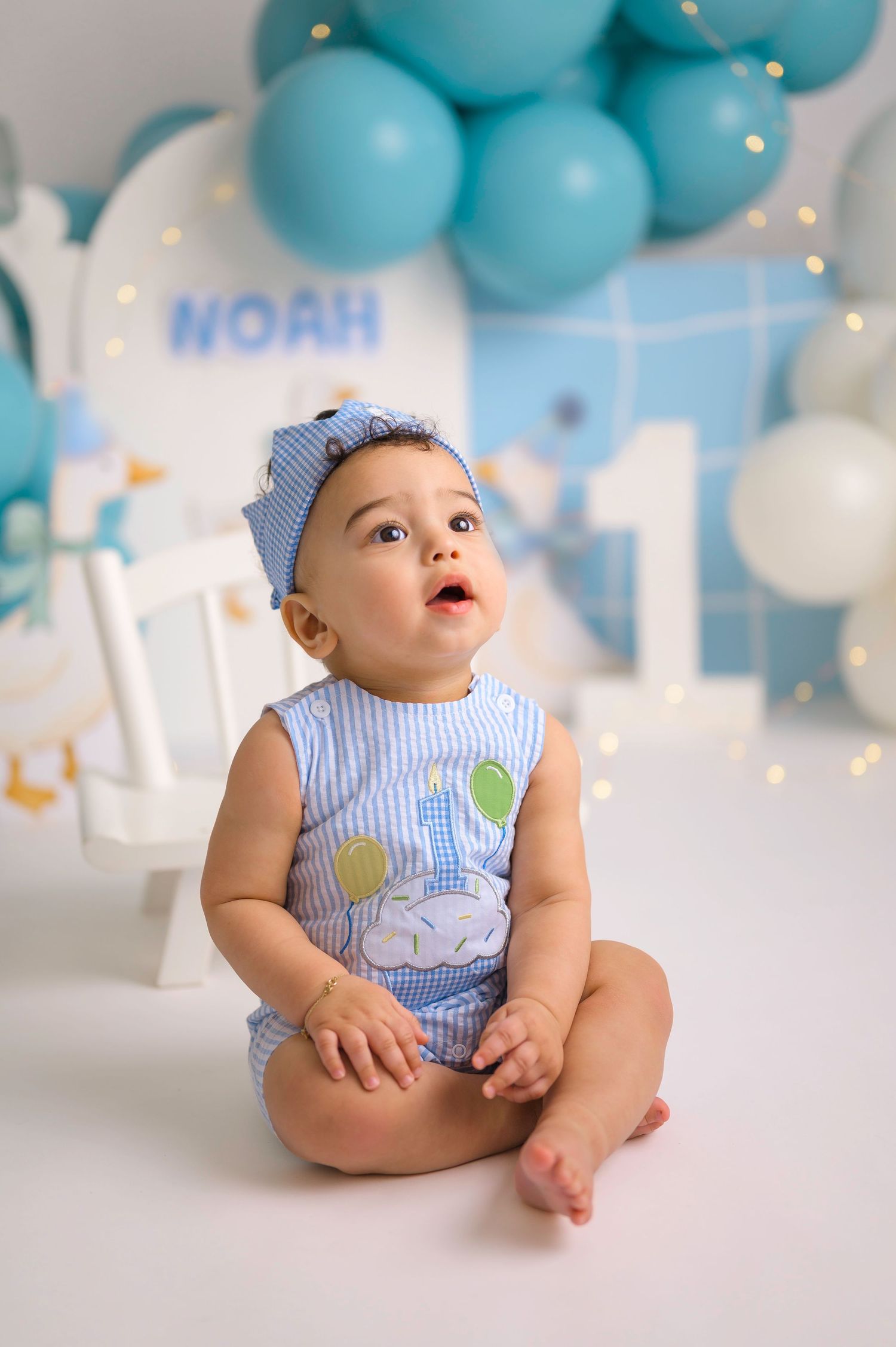 Baby Noah sits on white floor in blue outfit, celebrating his 1st birthday with balloons and decorations.