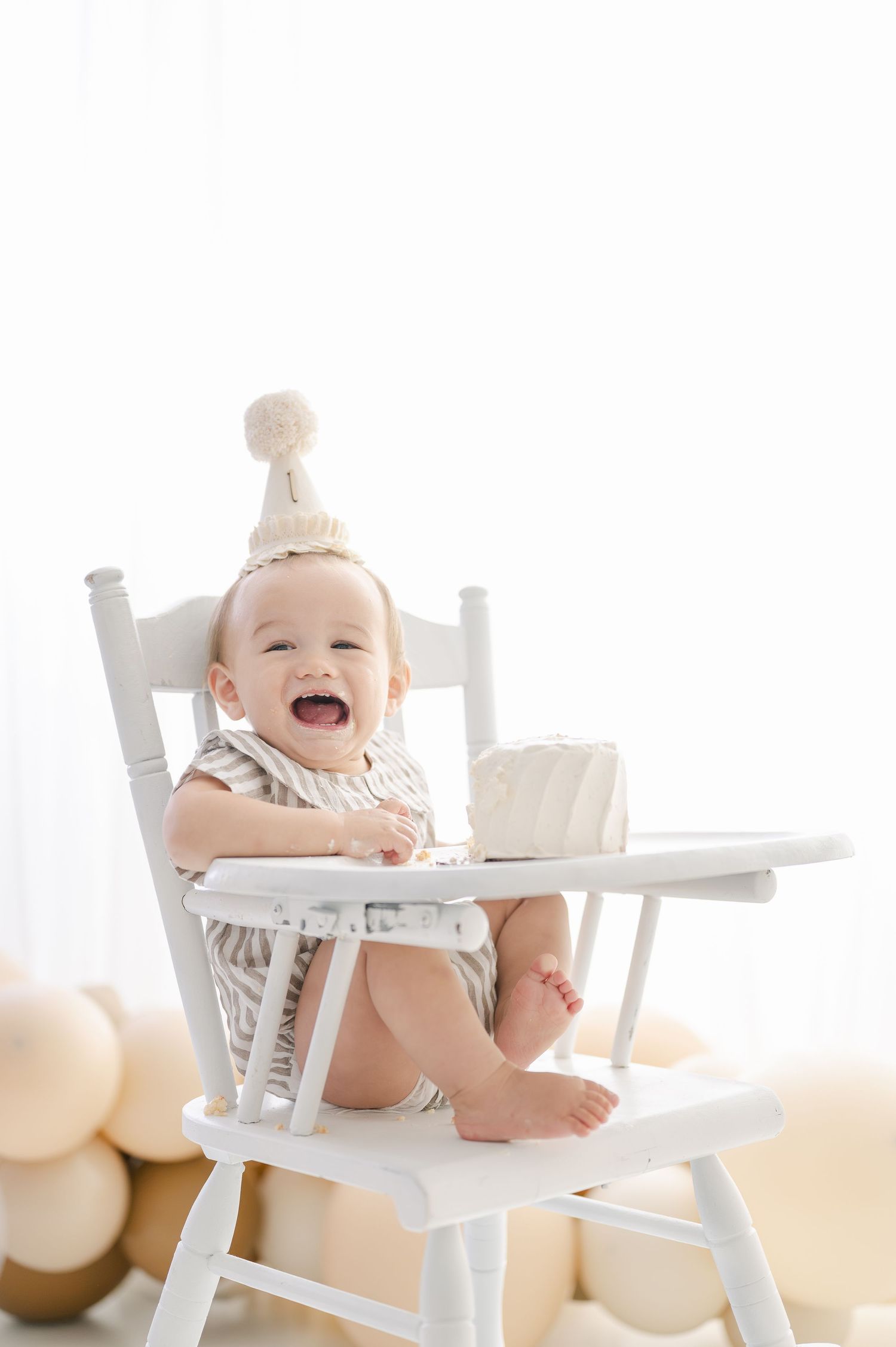 Happy baby in white high chair wearing a knit birthday hat, laughing during first birthday cake smash photo session.