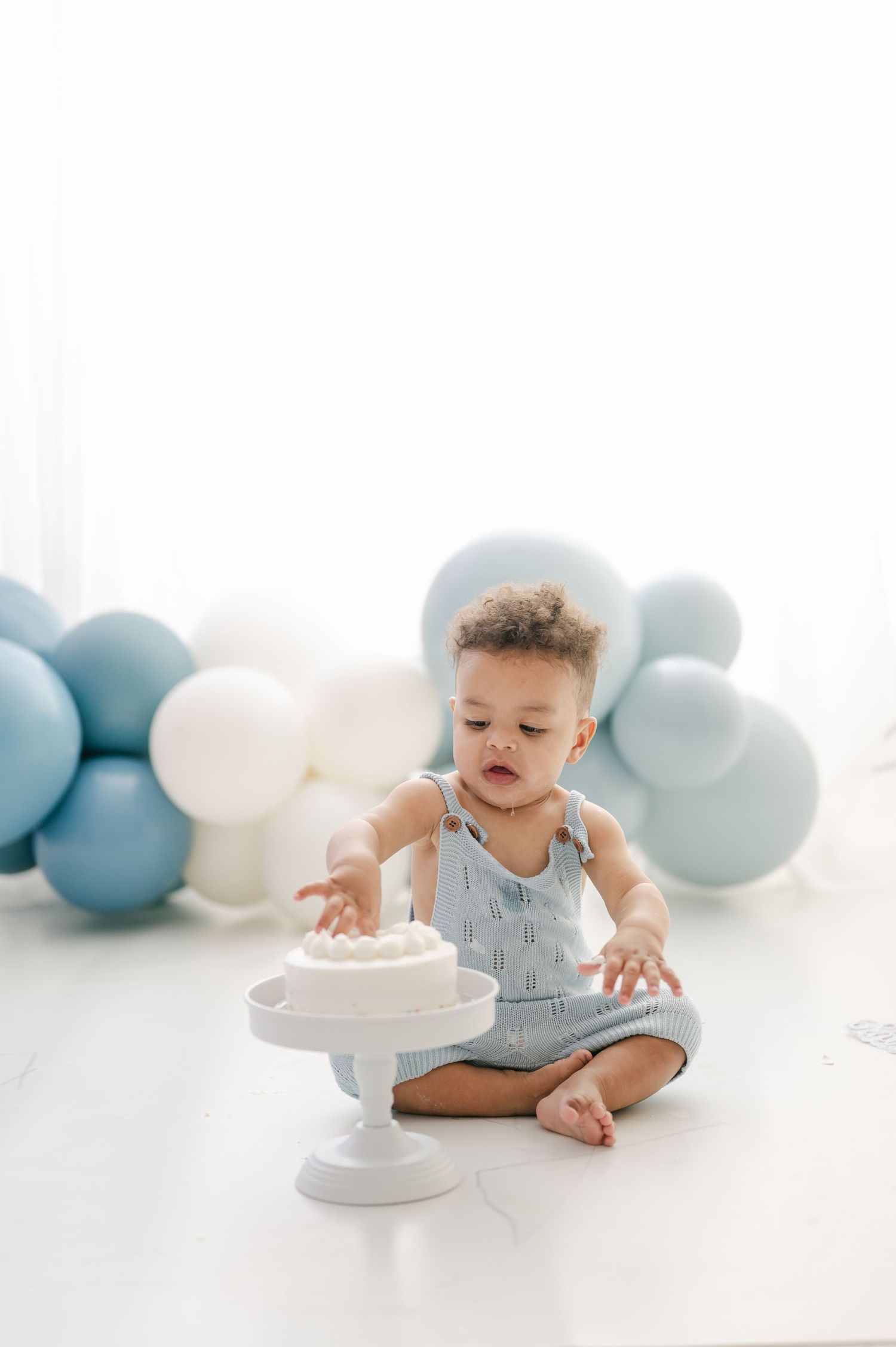 Baby boy in blue romper reaching for white smash cake surrounded by blue and white balloons on first birthday.