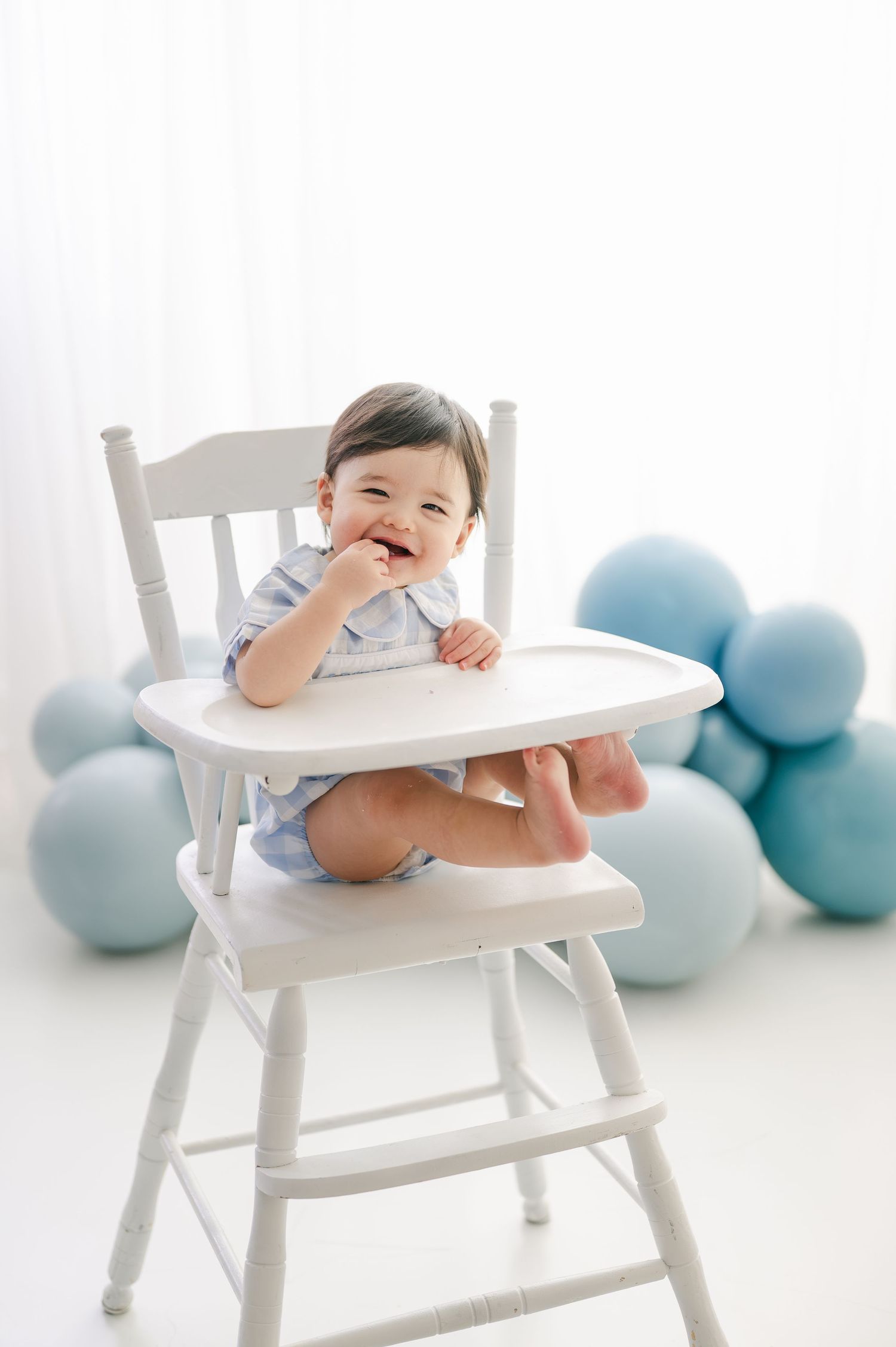Smiling baby in white high chair surrounded by blue balloons during first birthday cake smash photo session.