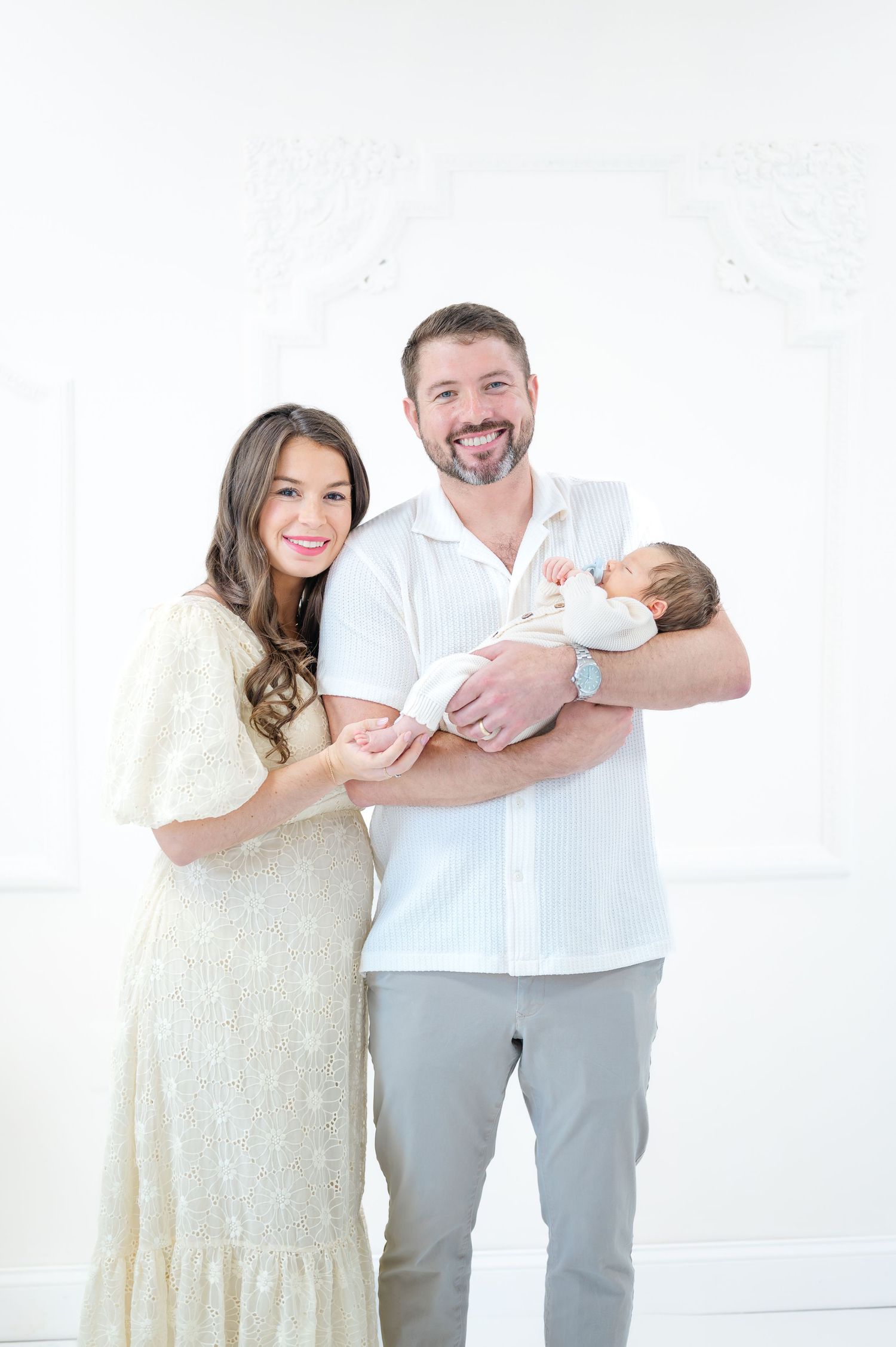 Smiling couple in white outfits holding their newborn baby in a bright, elegant studio setting.
