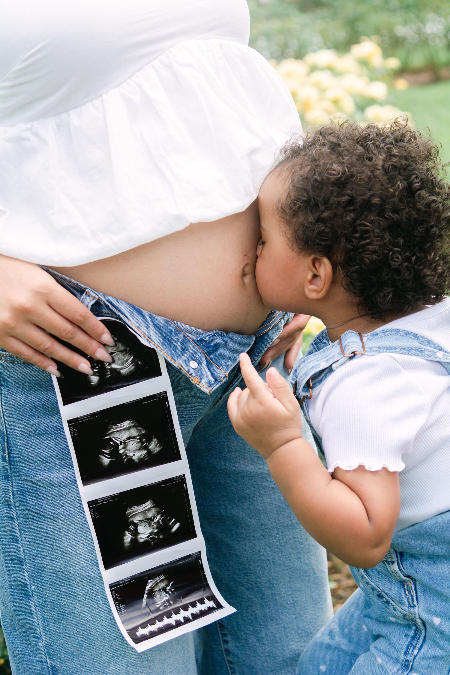 Child kisses pregnant belly while holding ultrasound photos outdoors.