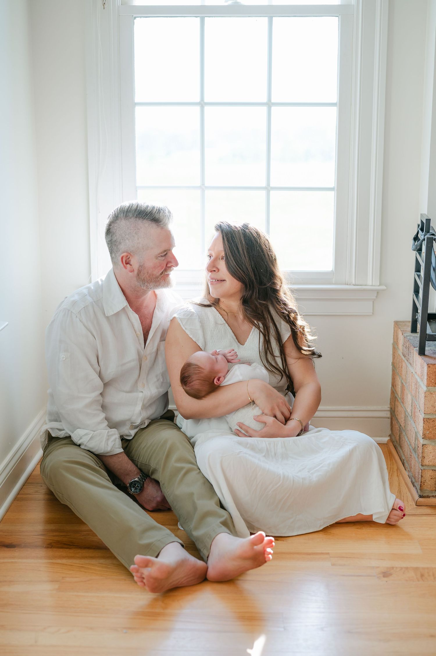 A couple sits barefoot on hardwood floors by a bright window, smiling at each other while cradling their newborn baby.