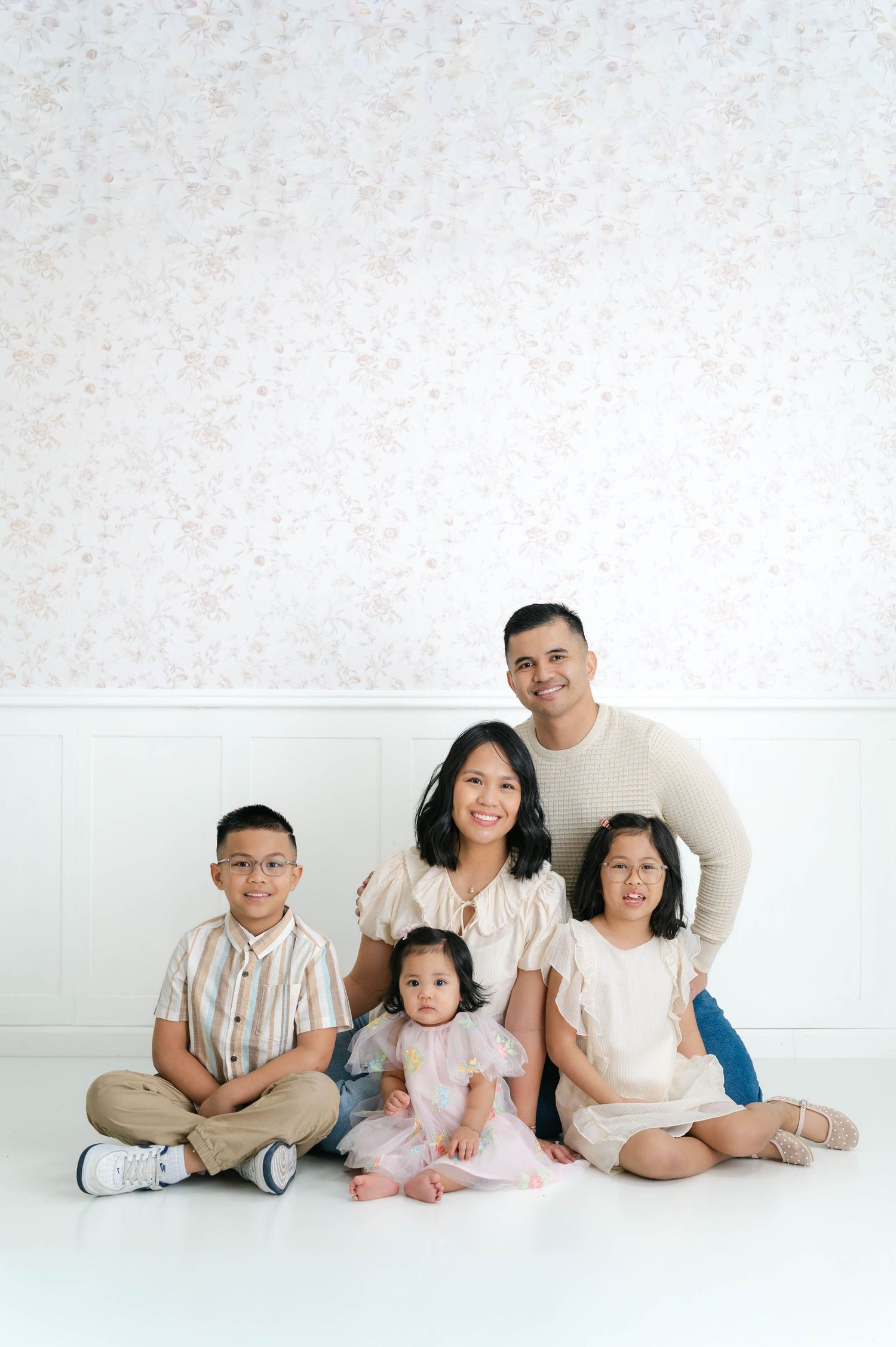 Family sitting together on white floor wearing neutral colored clothing in minimalist studio setting.