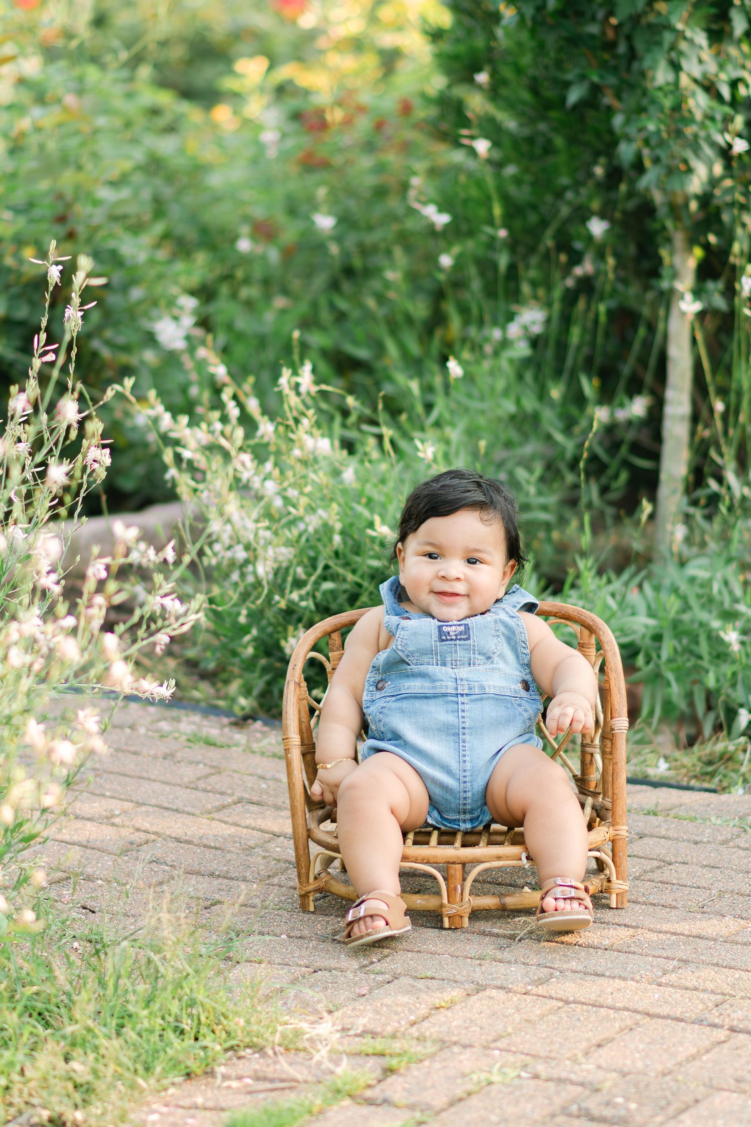 Baby in denim overalls sits in small wooden chair outdoors in garden setting.