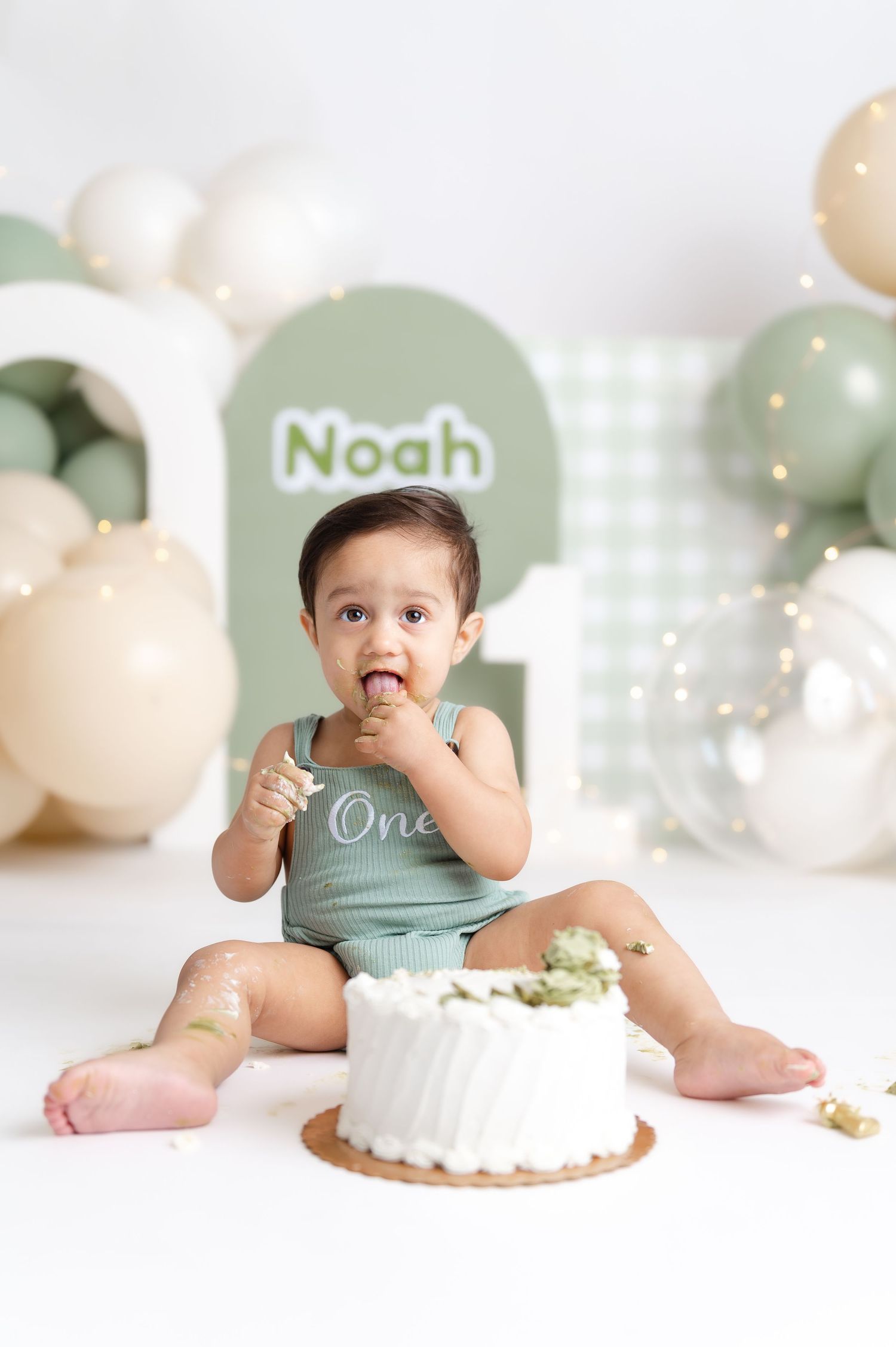 Baby Noah enjoys his first birthday cake smash, surrounded by sage green and neutral balloon decorations.