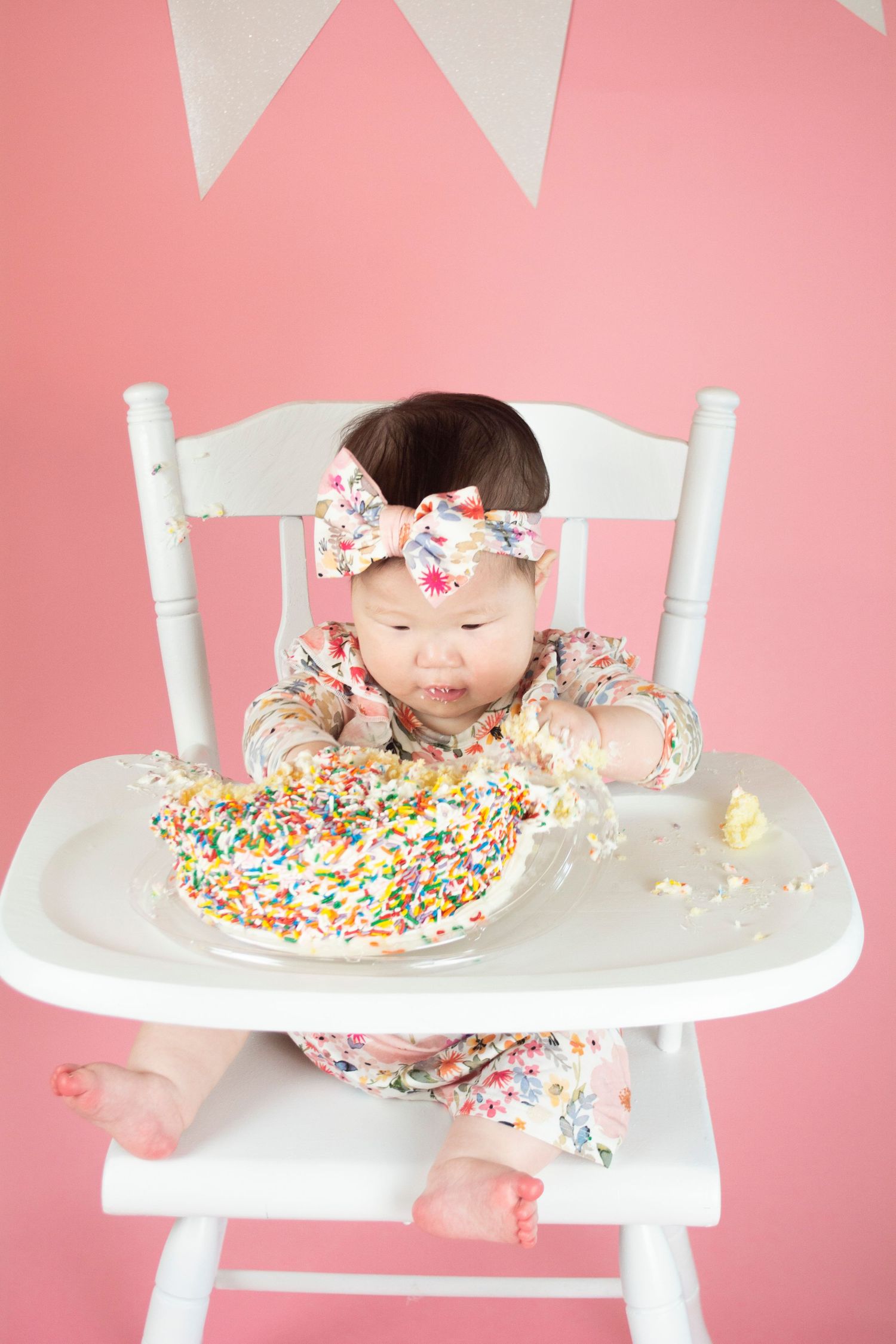 Baby leans forward in high chair to taste birthday cake with sprinkles against pink banner backdrop.