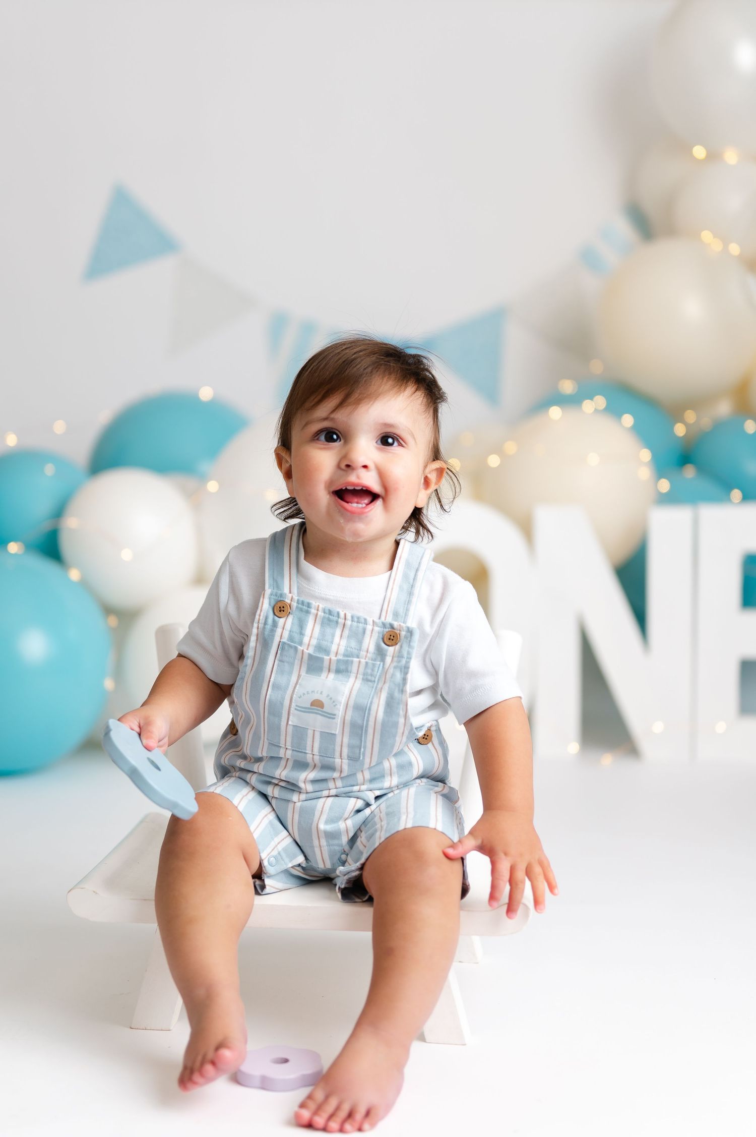 Happy baby boy in striped overalls sitting at his first birthday party with blue and white balloons.