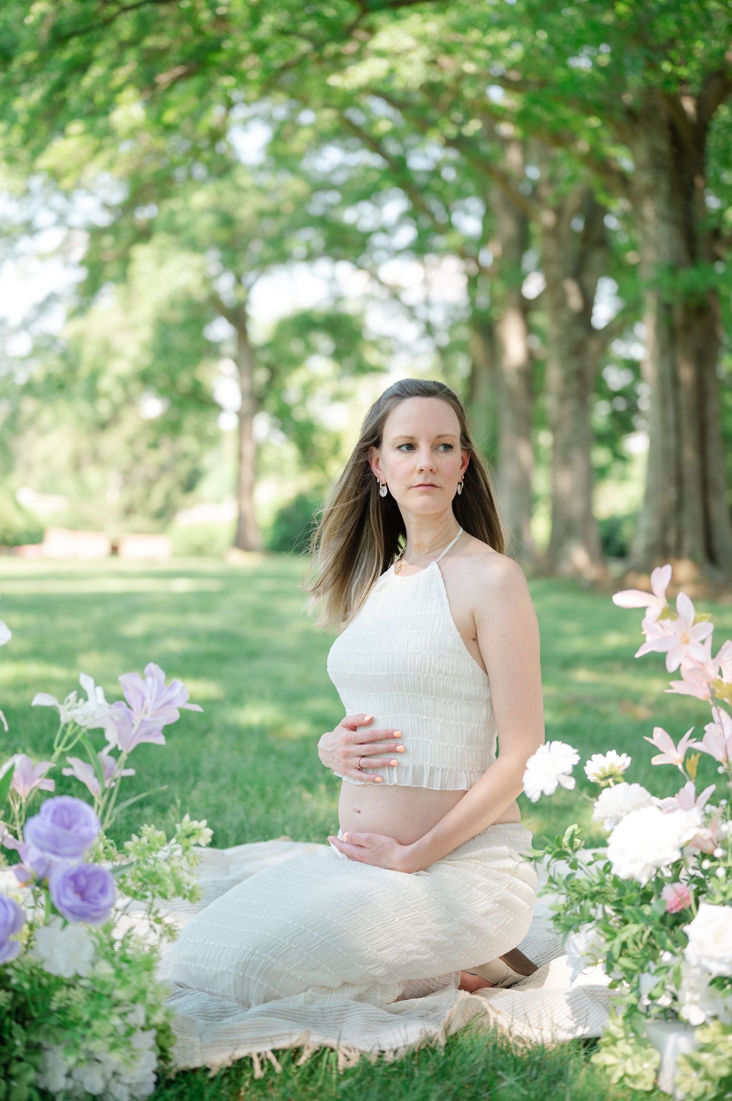 Pregnant woman in white crop top and tulle skirt sits among purple and white flowers in a lush green park.