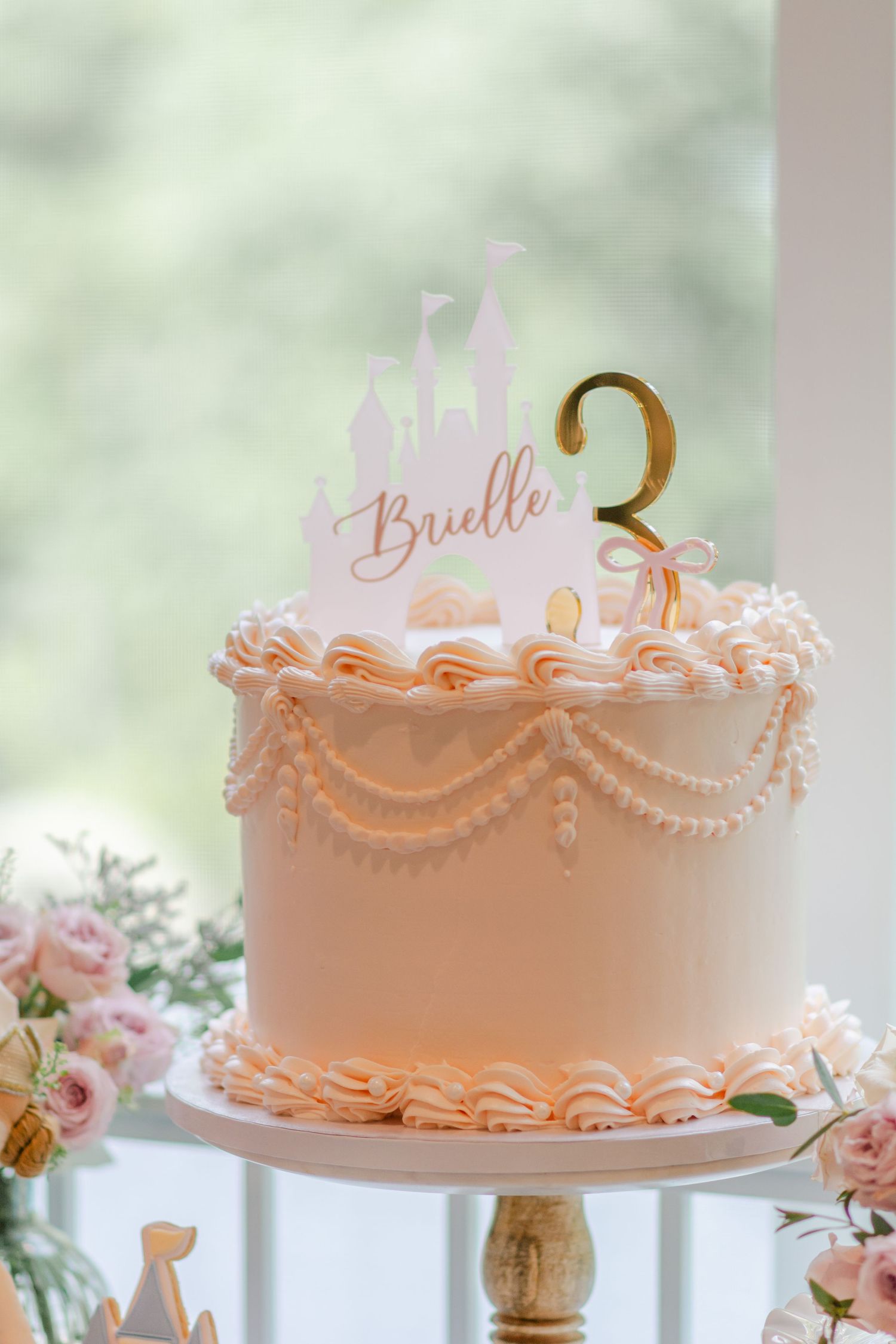 Pink frosted celebration cake with delicate piped details and pearl accents displayed on a gold cake stand.
