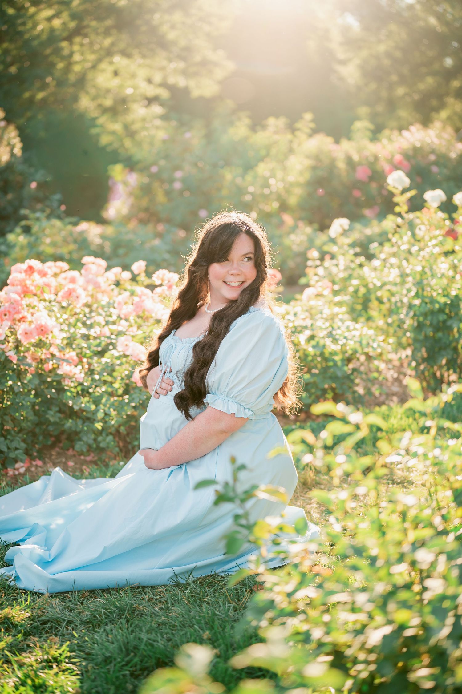 Pregnant woman in light blue gown sitting among pink roses in golden sunlight, smiling warmly at camera.