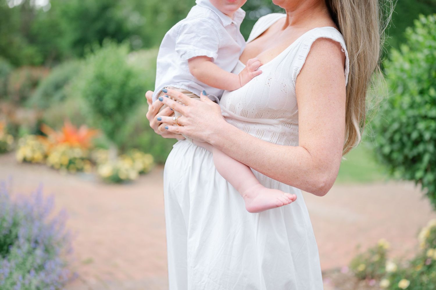 Pregnant woman in white dress holding baby in lush garden setting.
