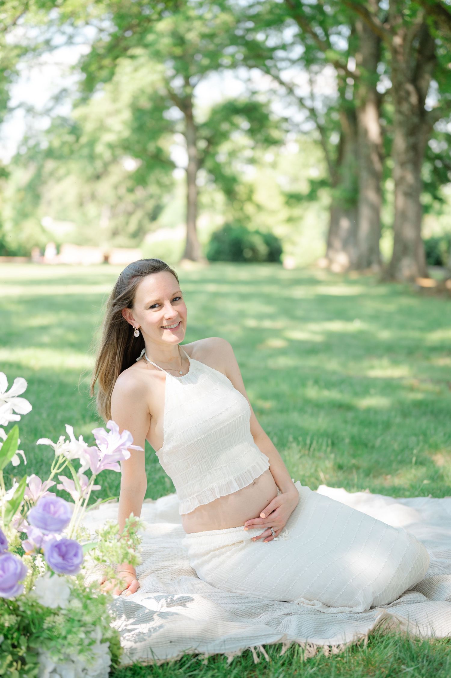 Pregnant woman in white crop top and skirt sits on blanket in lush green park surrounded by purple flowers.